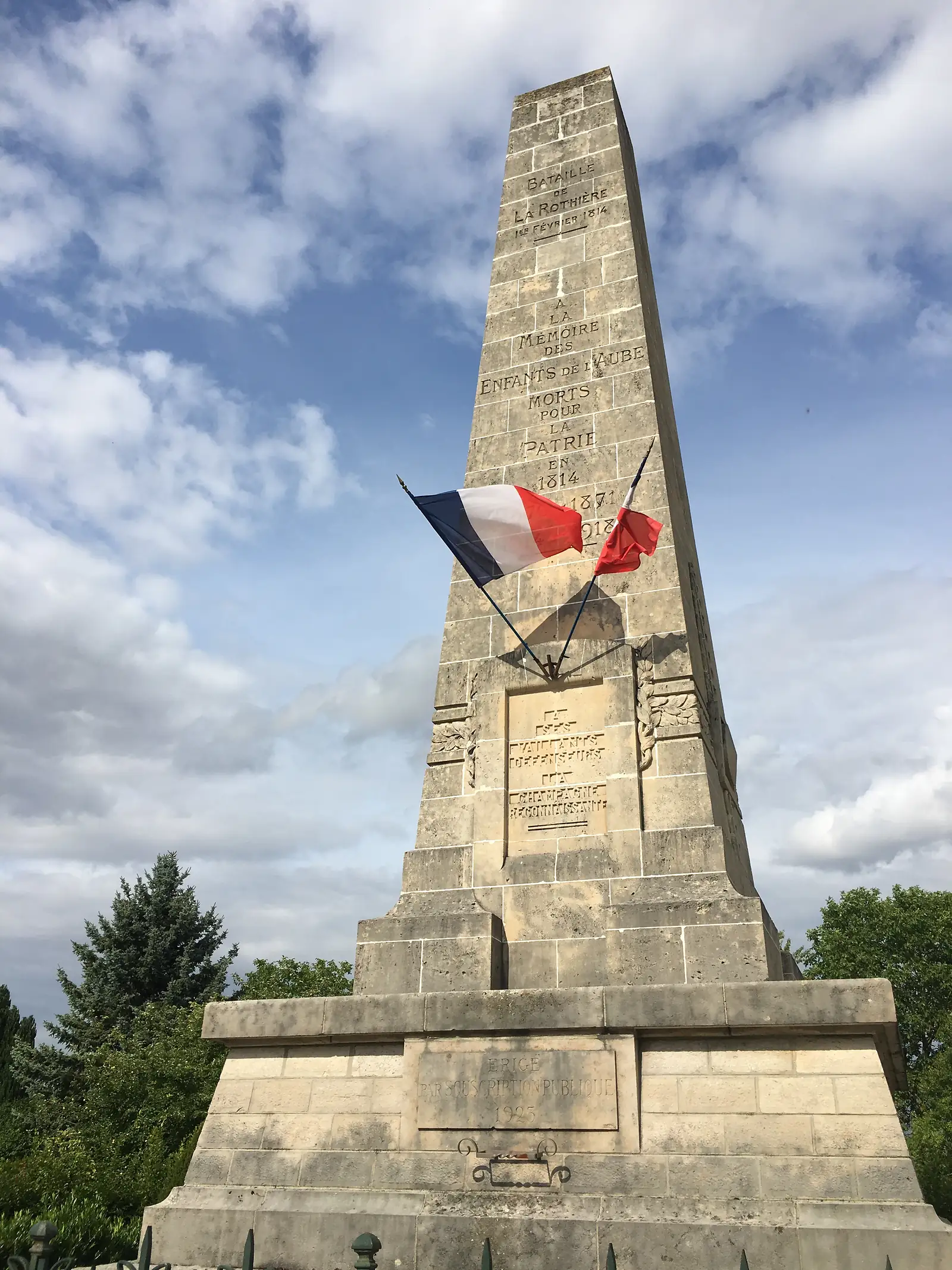 A stone war memorial obelisk at La Rothière, inscribed BATAILLE DE LA ROTHIÈRE 1er FÉVRIER 1814 and dedicated to the commune's dead of 1814, 1871 and 1914-1918, flying French tricolour flags.