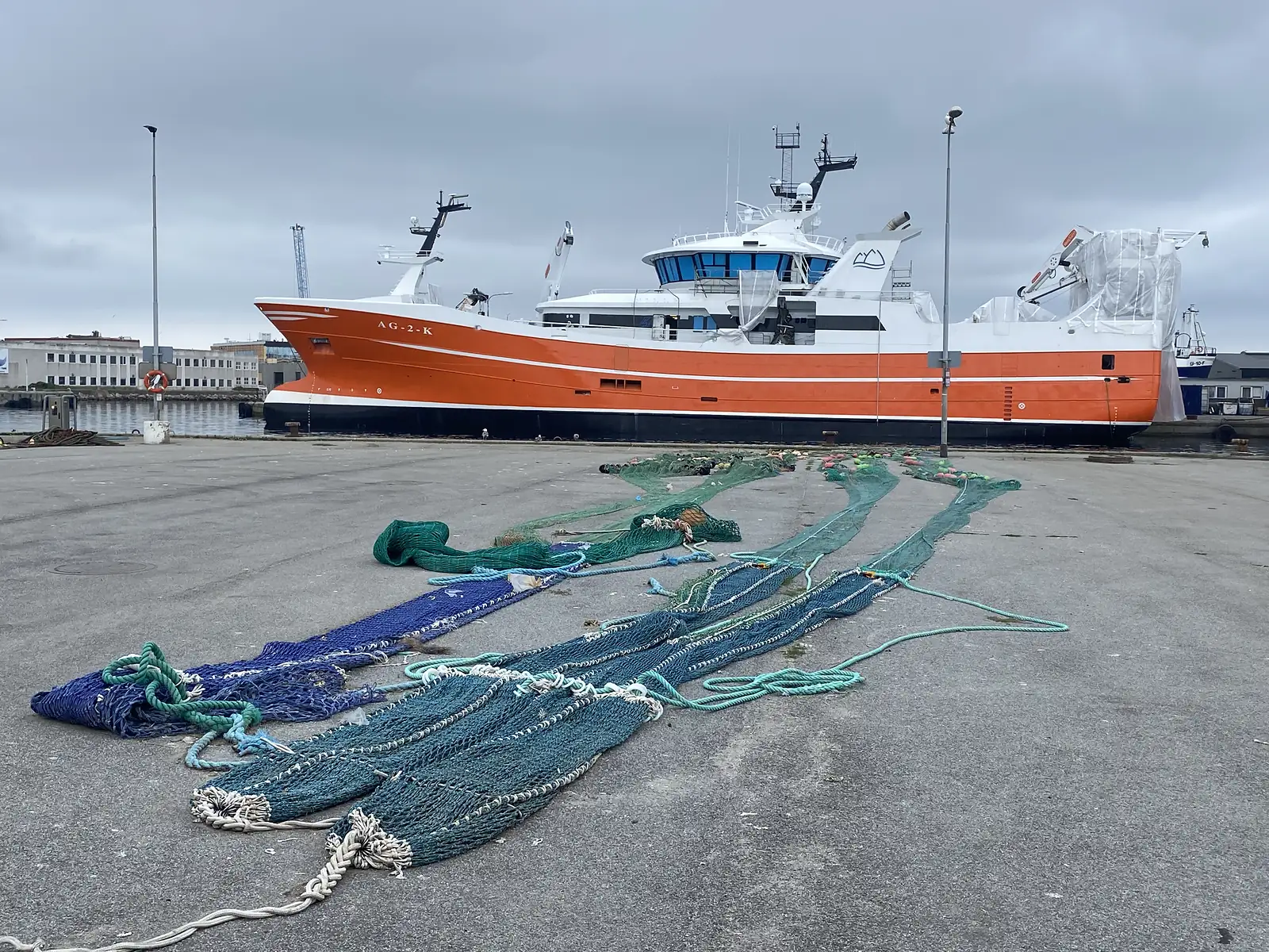 A large orange-and-white fishing trawler lettered "AG-2-K" moored at a concrete quay, with long green and blue trawl nets laid out on the dockside.