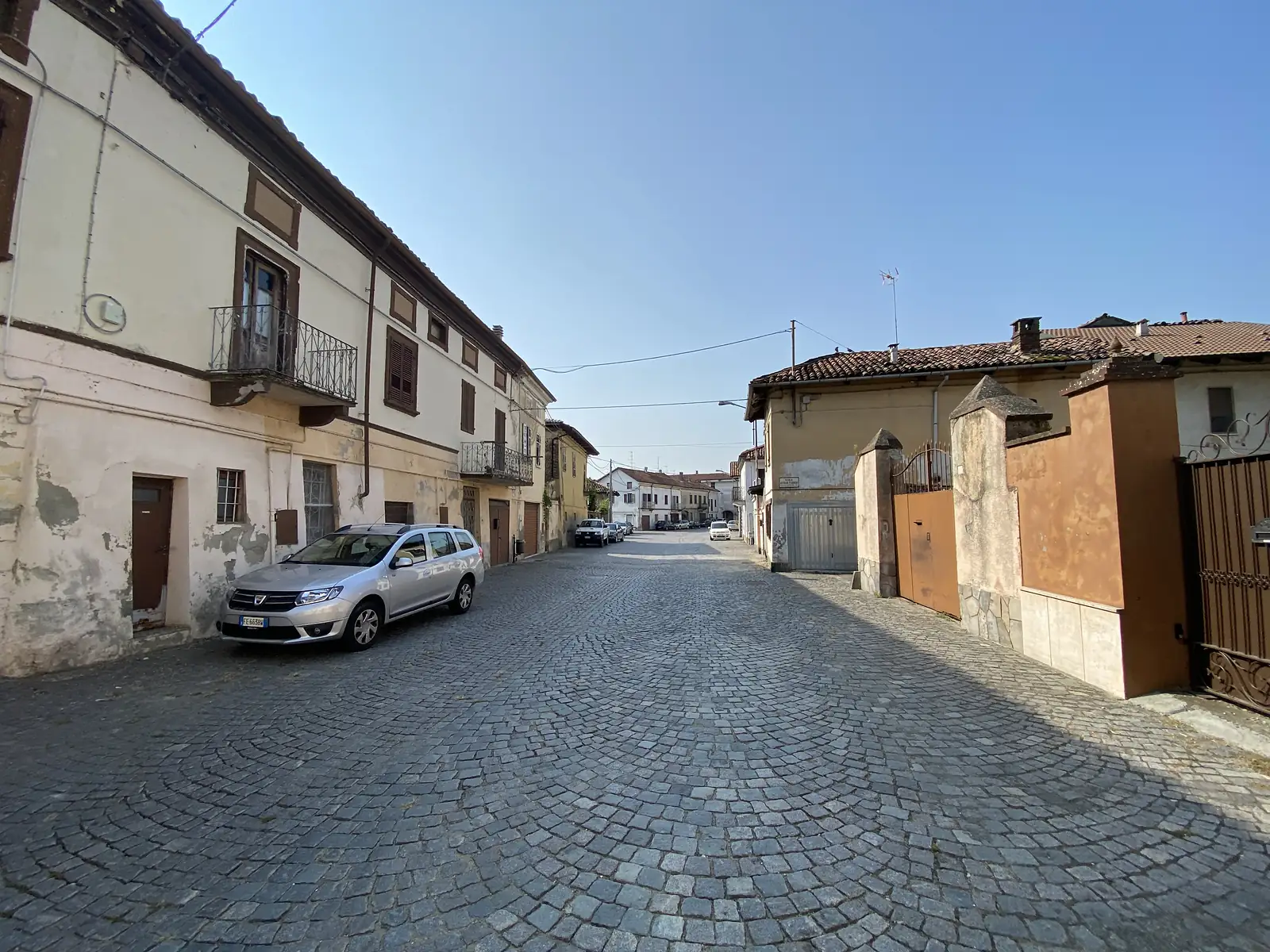 A quiet cobbled street between weathered plastered houses, with a silver estate car parked outside a faded doorway.