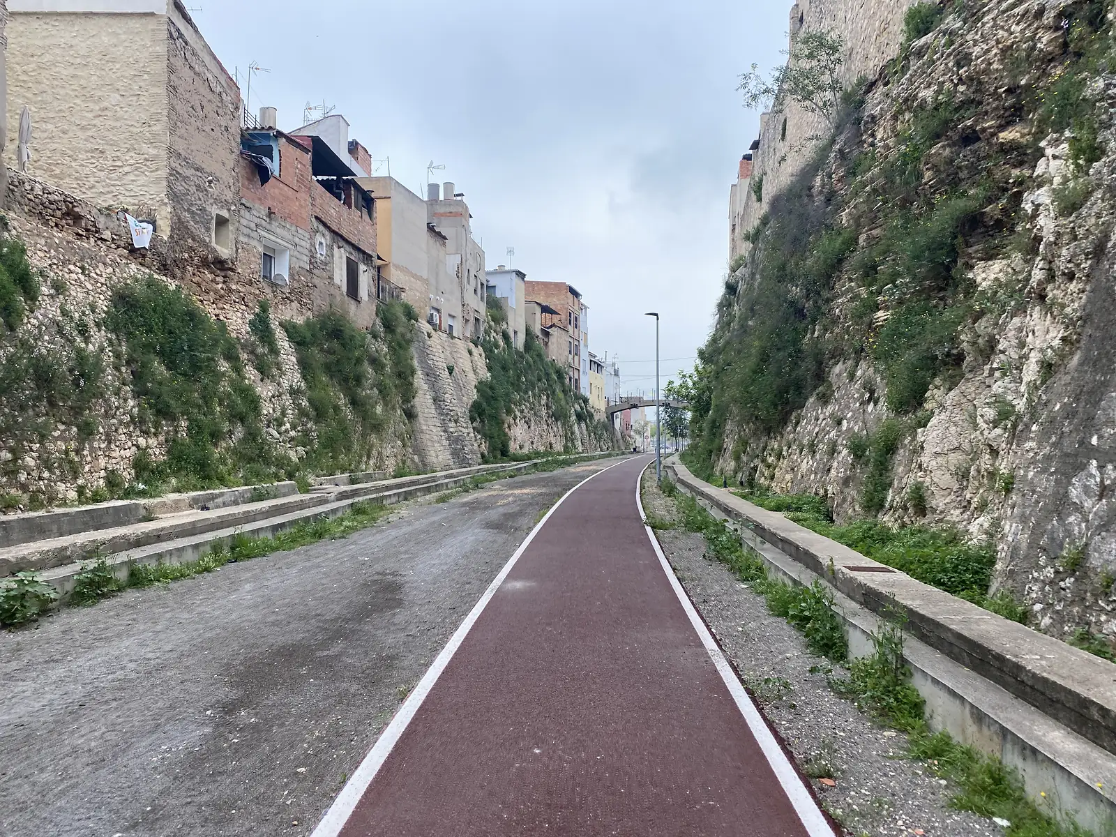 A red-surfaced cycle path running along the floor of a rock cutting through a town, houses perched on the retaining wall above.