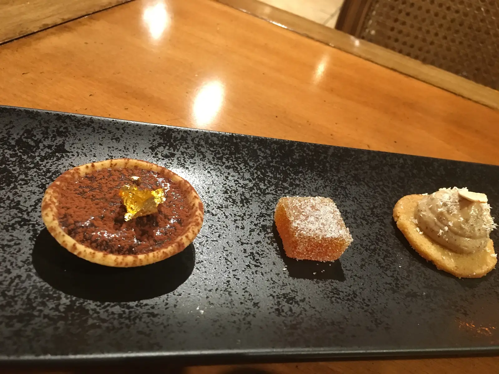 Three petits fours on a dark tray: a cocoa-dusted chocolate tartlet with gold leaf, a sugar-coated fruit jelly, and a spiced cream on a biscuit base.