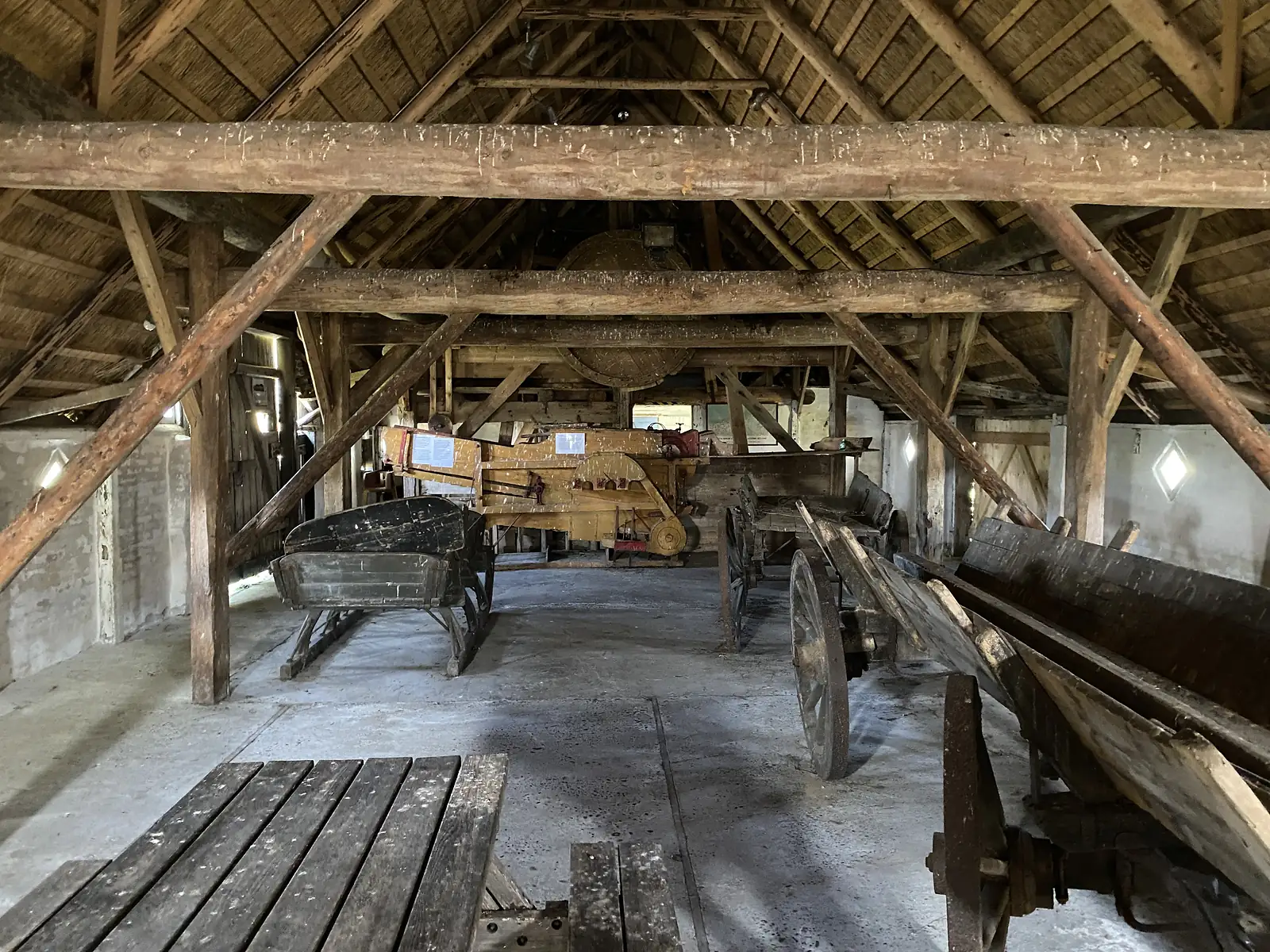 The interior of a thatched timber-framed barn with heavy roof beams, old horse carts, a wagon and farm machinery standing on the floor.