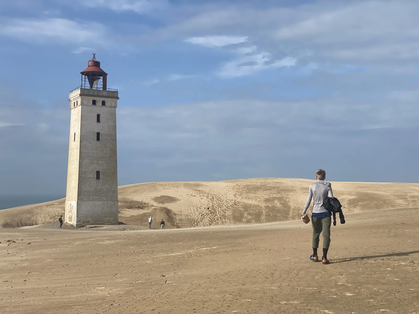 A tall white lighthouse with a red lantern standing on bare sand, with people walking across the dune towards it.