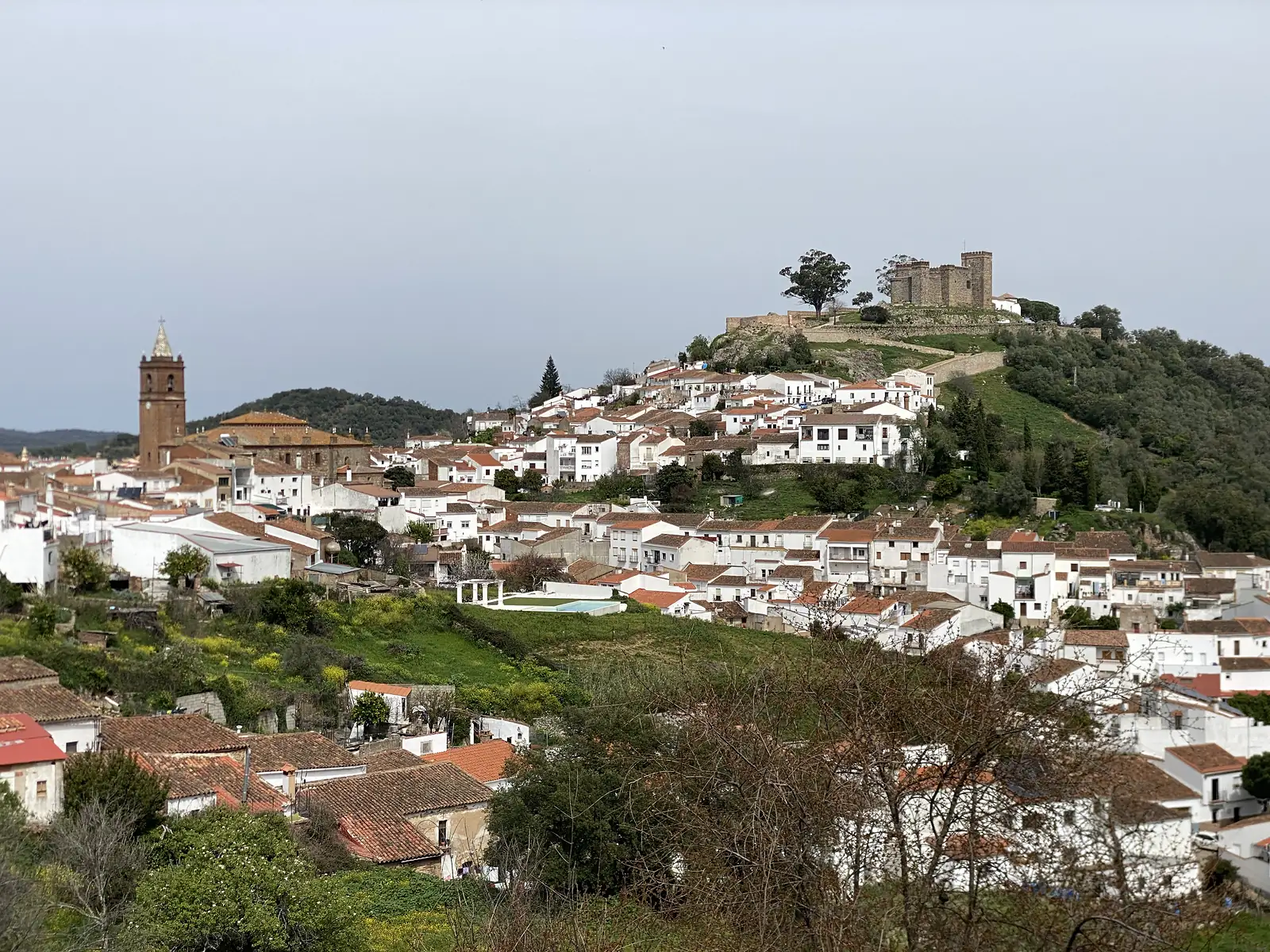 A whitewashed hill town with a tall brick bell tower at its heart and a long castle wall running along the green hilltop above it.