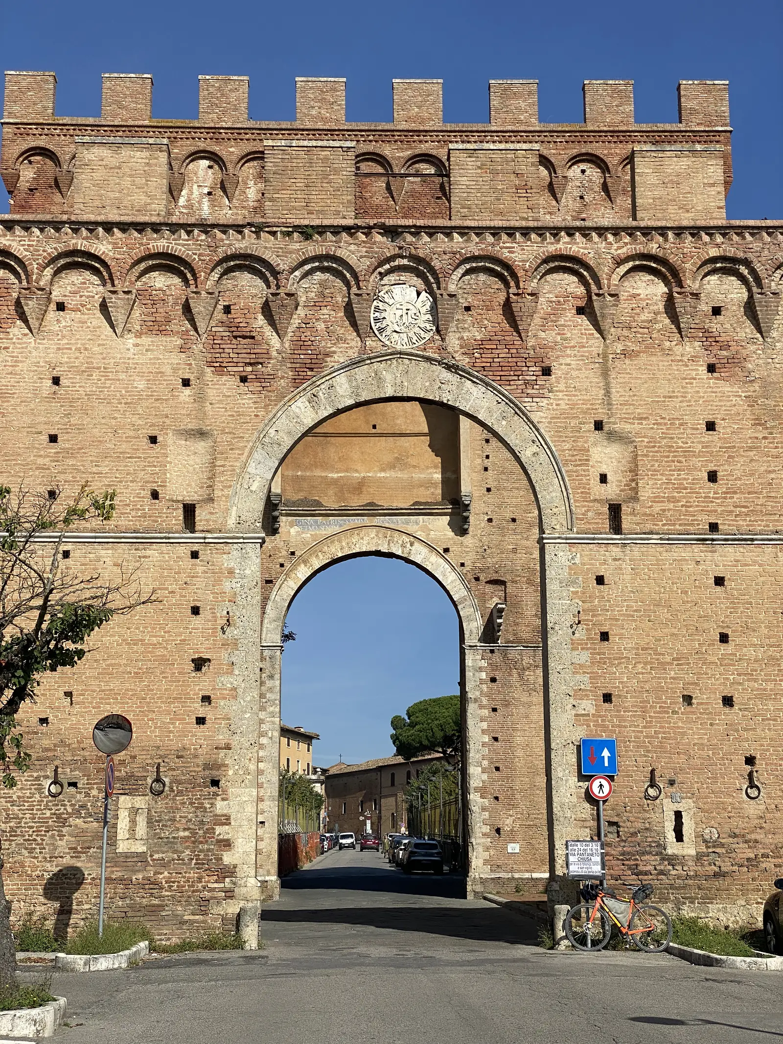 A medieval brick city gate with a crenellated top and a tall arched passage through the wall, with a laden bike parked beside it.
