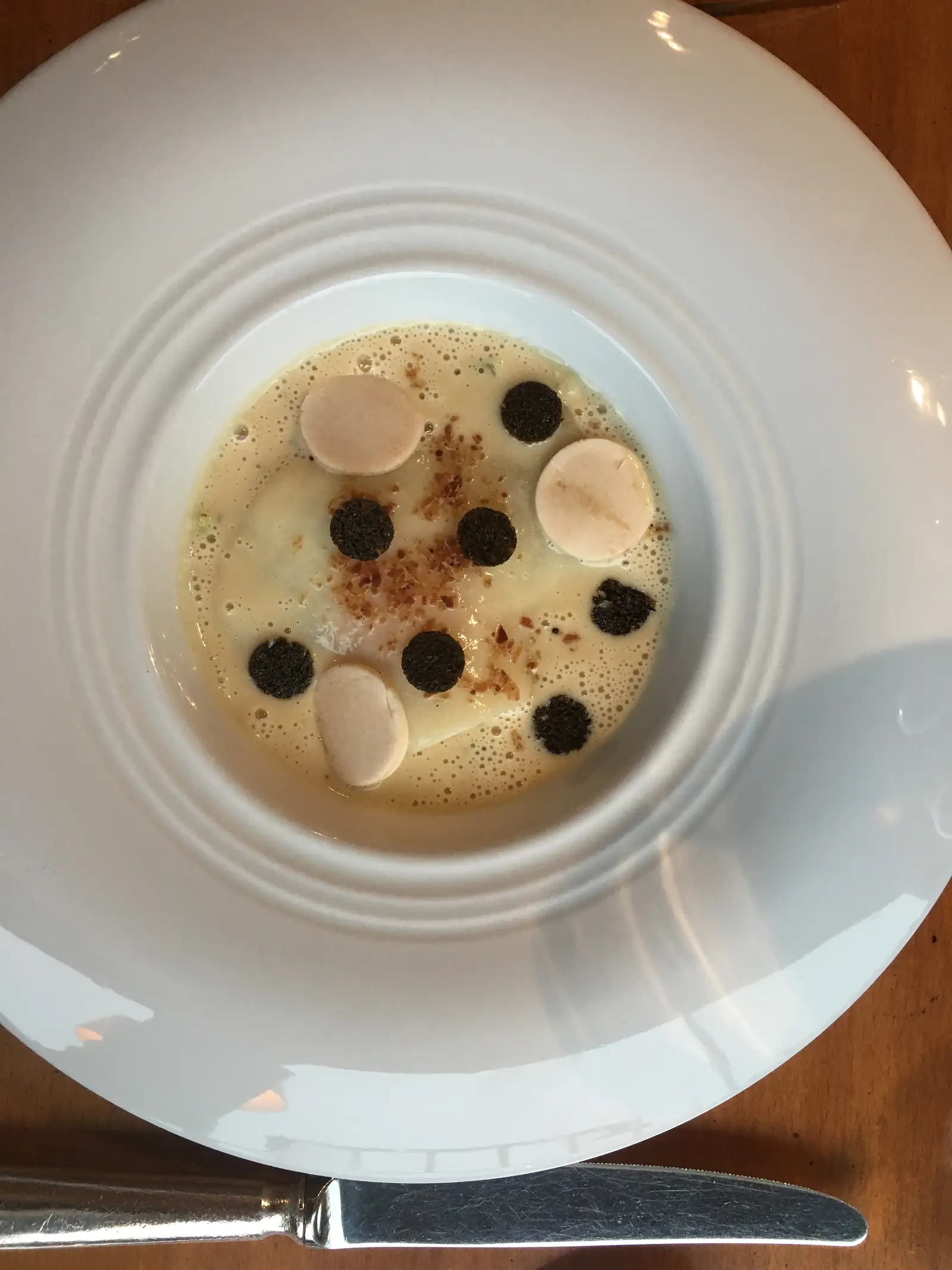 A fine-dining dish of pale discs and dark truffle rounds arranged over a light, frothy sauce in a wide white bowl.