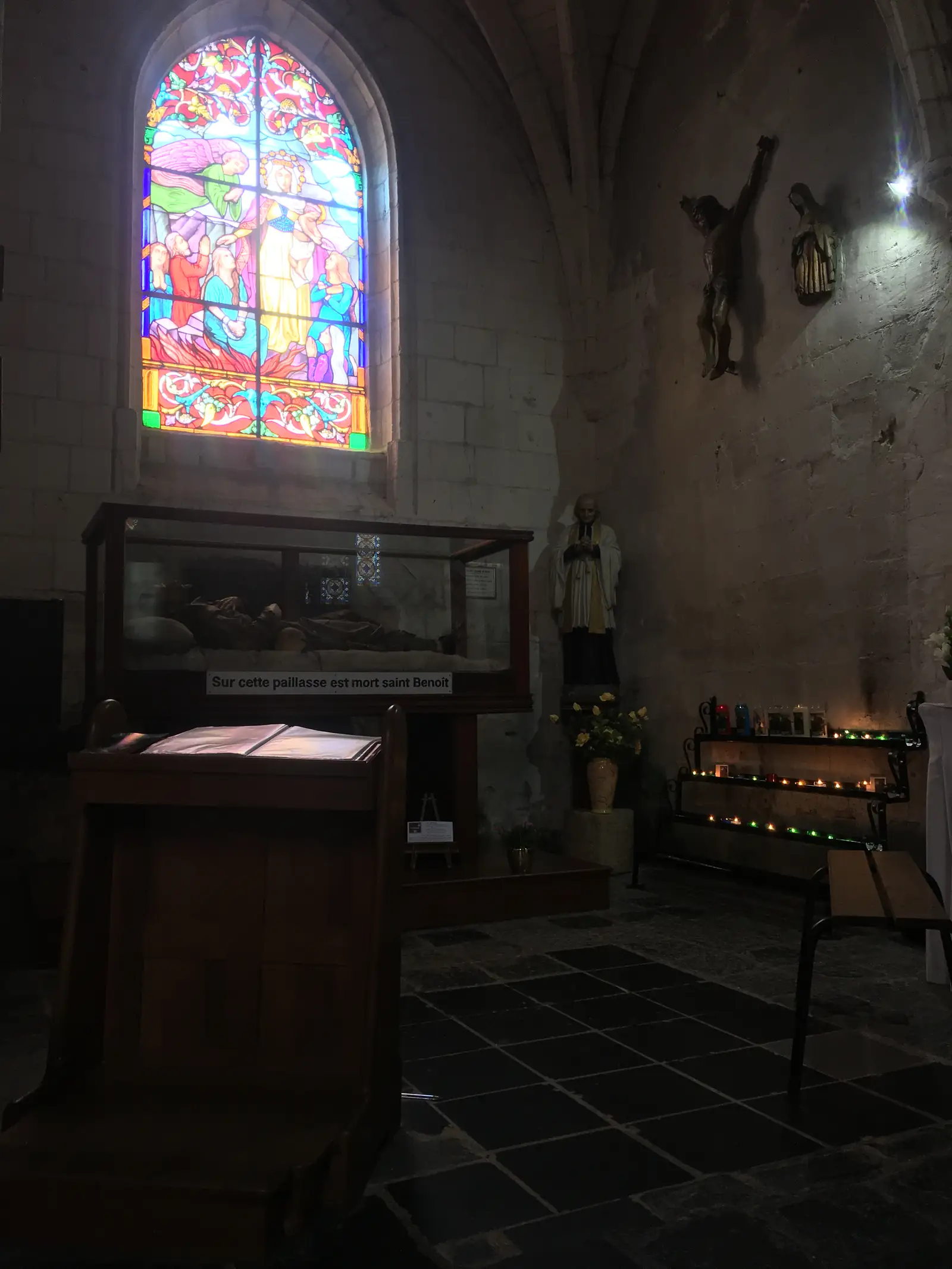 Inside a church chapel, a glass case beside a carved crucifix and a colourful stained-glass window bears a sign reading 'Sur cette paillasse est mort saint Benoît'.