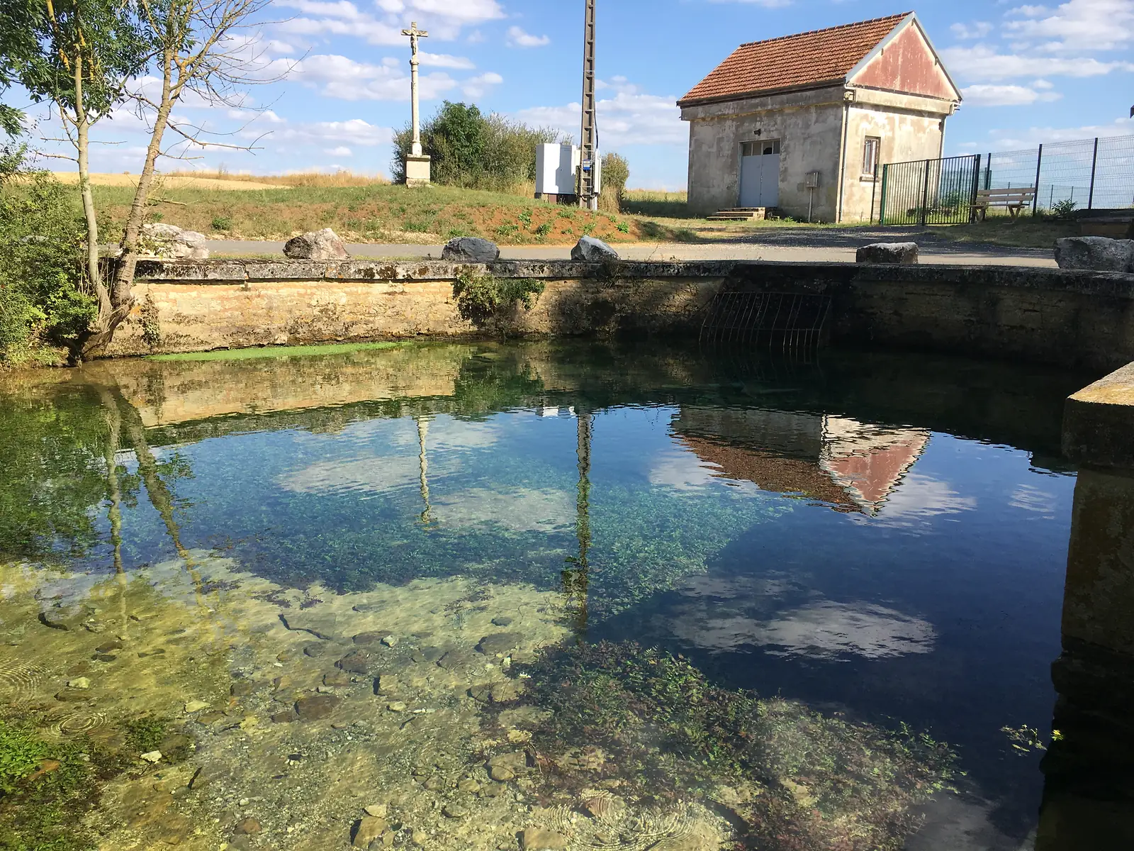 A clear, spring-fed pool reflects a small stone building and a roadside crucifix, with an iron grille set into the retaining wall.