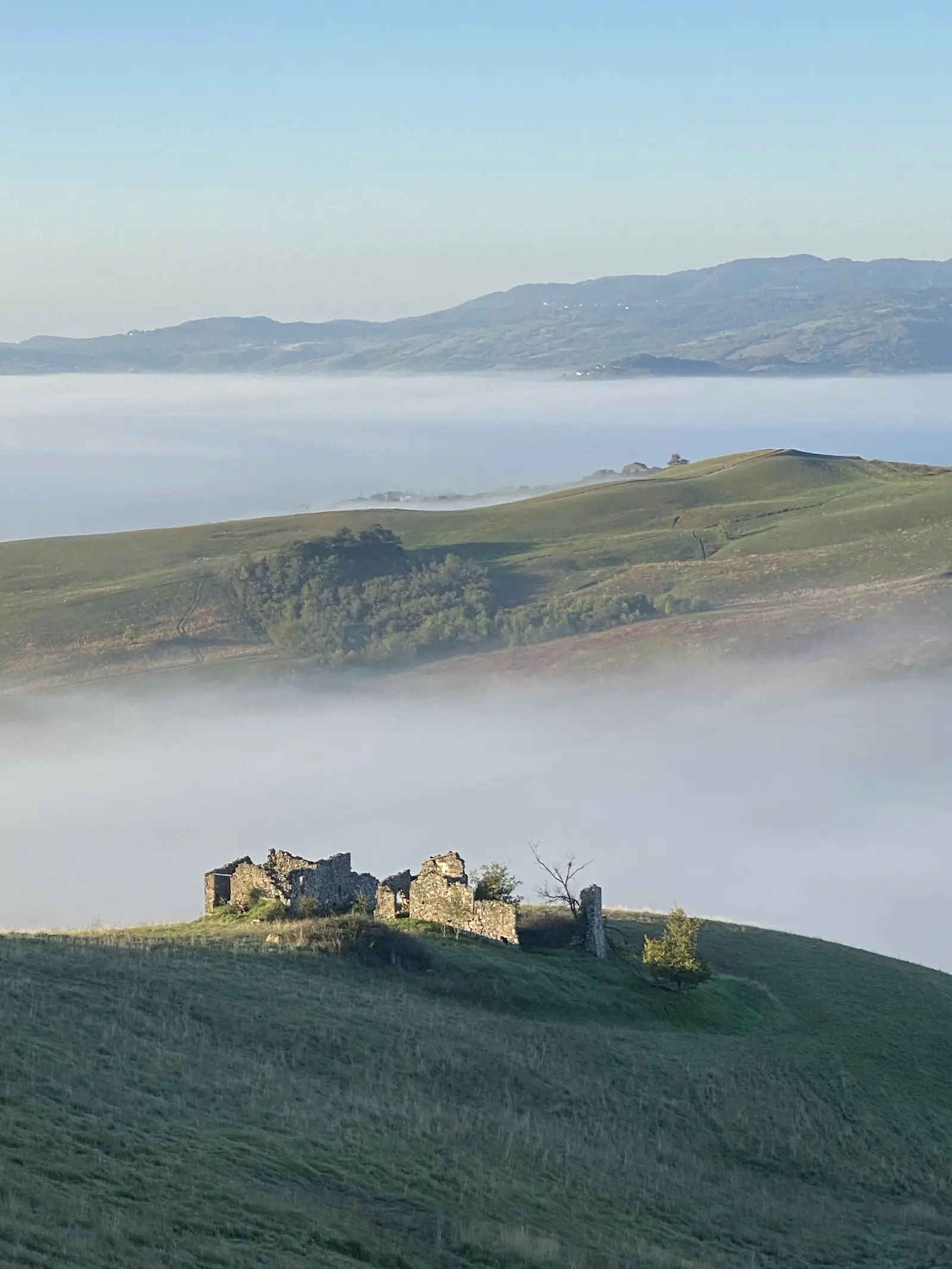A crumbling stone ruin on a grassy hilltop overlooking a valley filled with cloud, distant hills rising above the mist.
