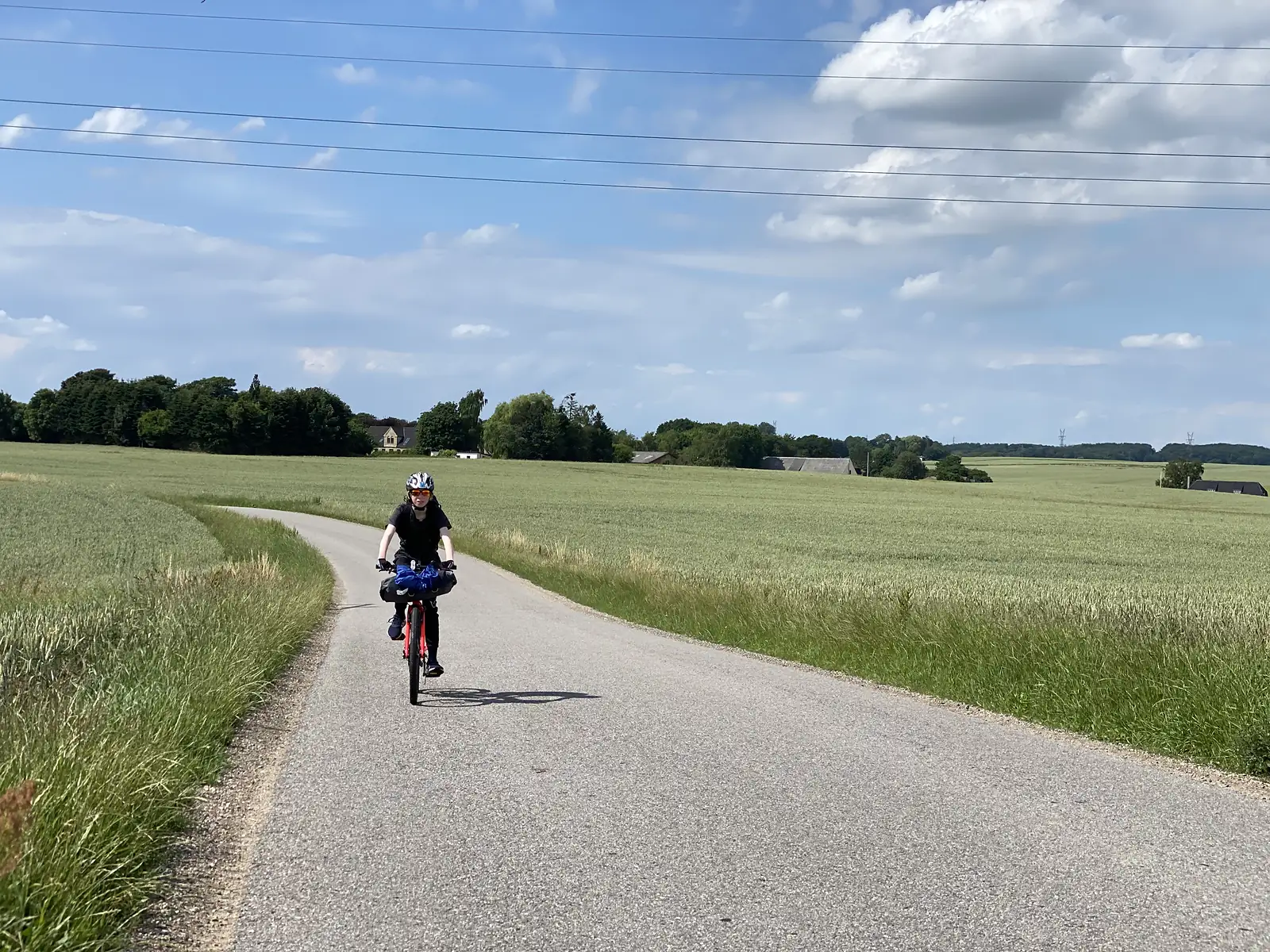 A young cyclist on a loaded red bike riding towards the camera along a narrow lane between green cereal fields, with farms and trees behind.