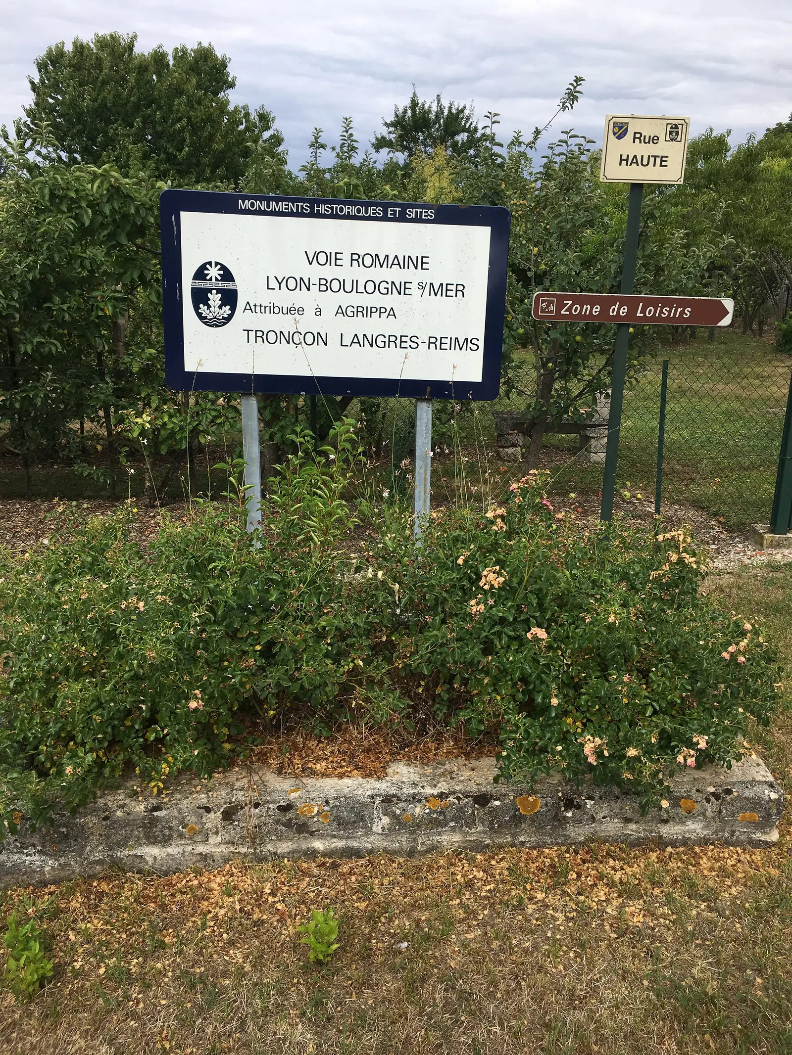 A heritage sign reading VOIE ROMAINE LYON-BOULOGNE S/MER, ATTRIBUÉE À AGRIPPA, TRONÇON LANGRES-REIMS stands among flowering shrubs near a garden fence.