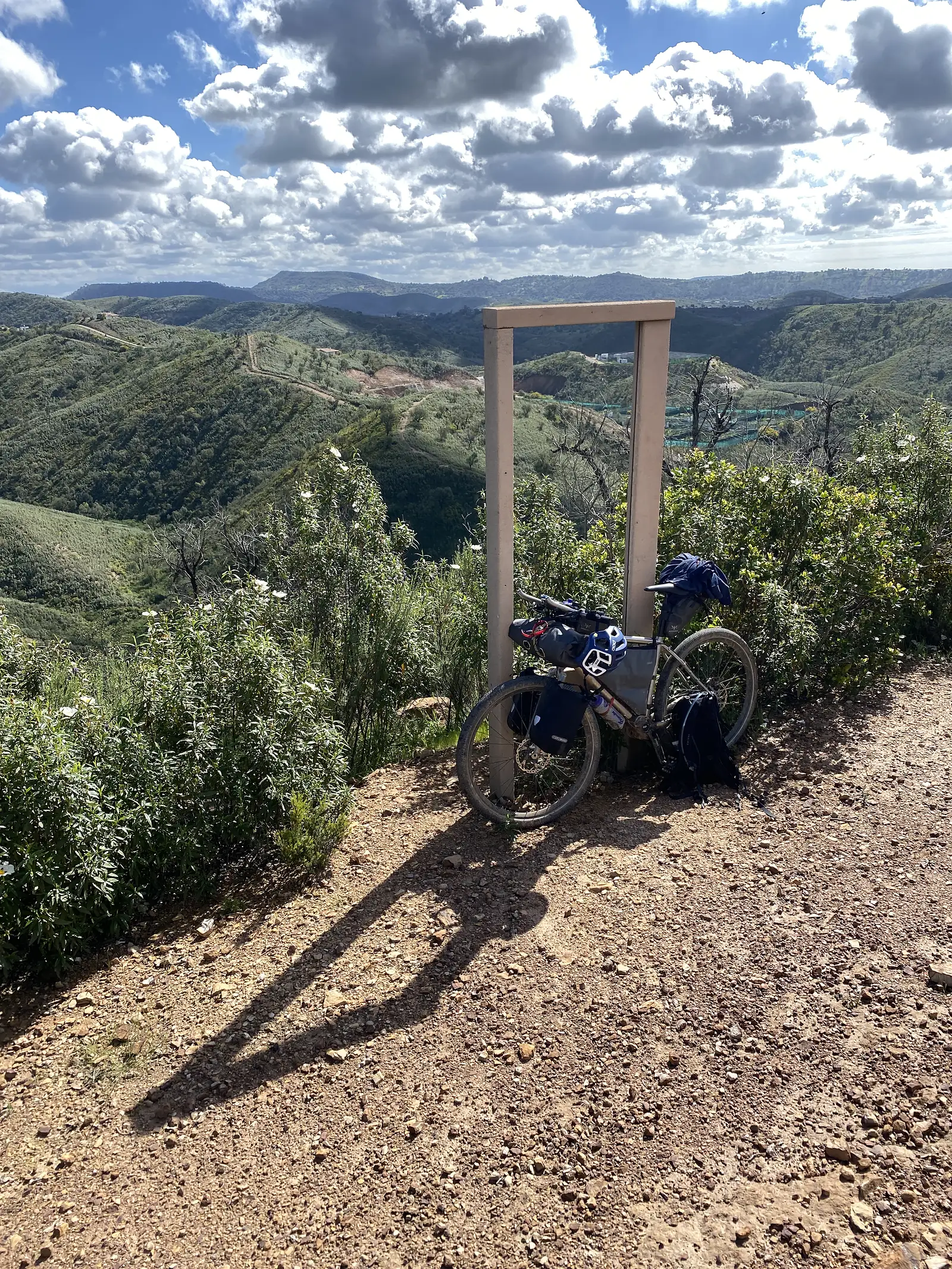 Loaded bike leaning against a plain concrete frame marking a roadside viewpoint, looking out over rolling green hills scored with tracks under a sky of broken cumulus.