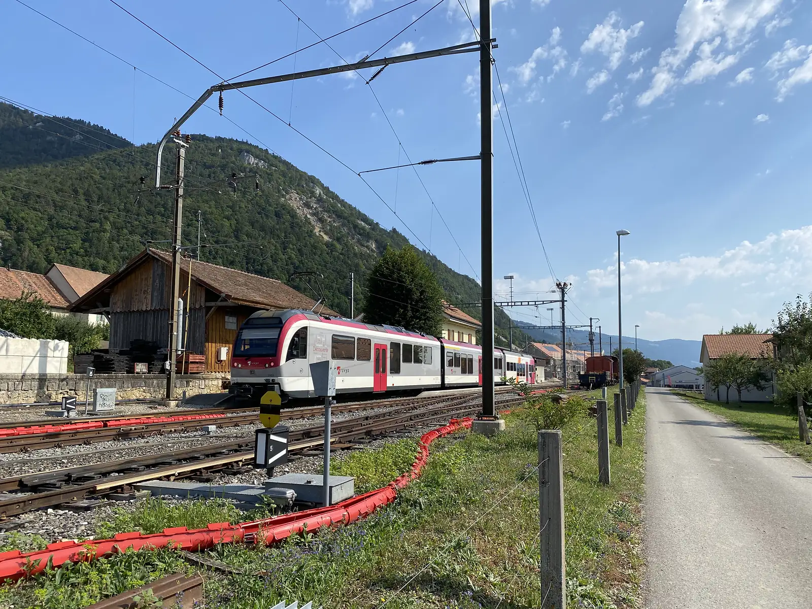 A pink-and-white Travys train arriving at a rural station, overhead wires crossing above the platform and a wooded hillside behind.