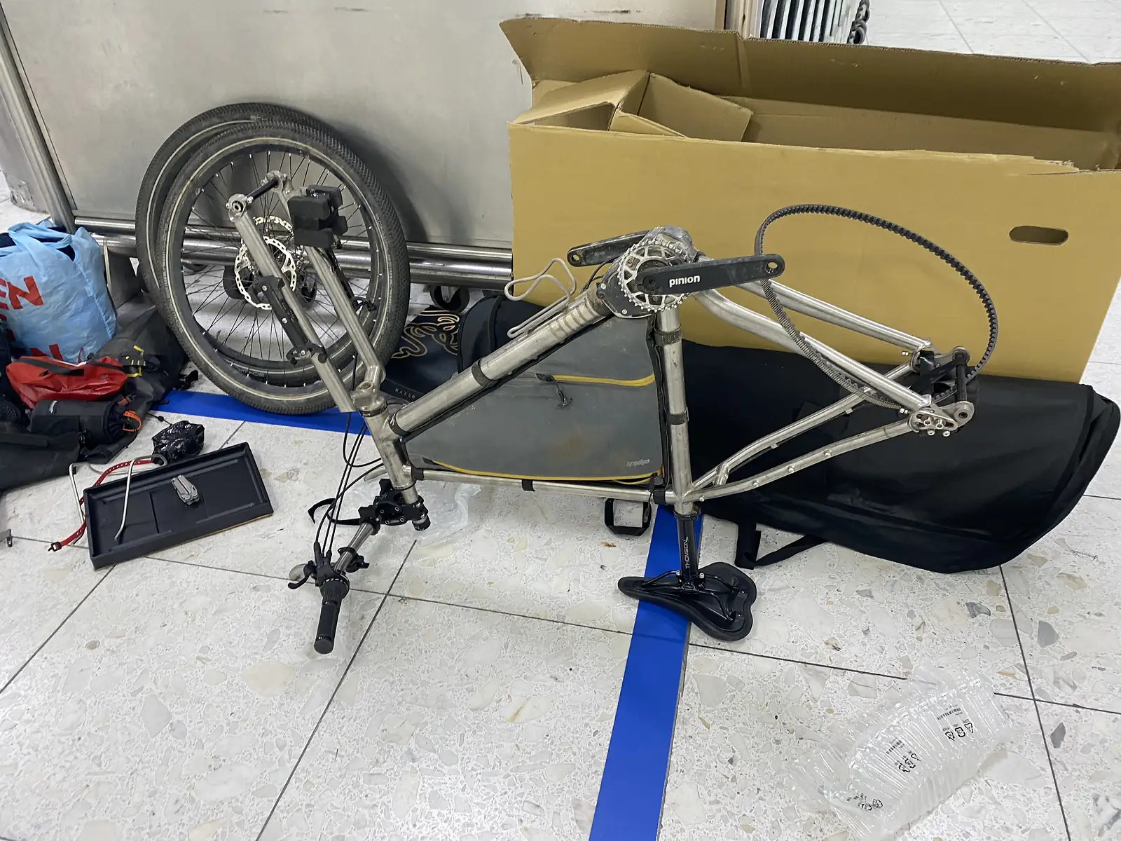 A titanium bike frame with belt drive standing part-built on an airport floor beside its wheel, cardboard box and travel bag.
