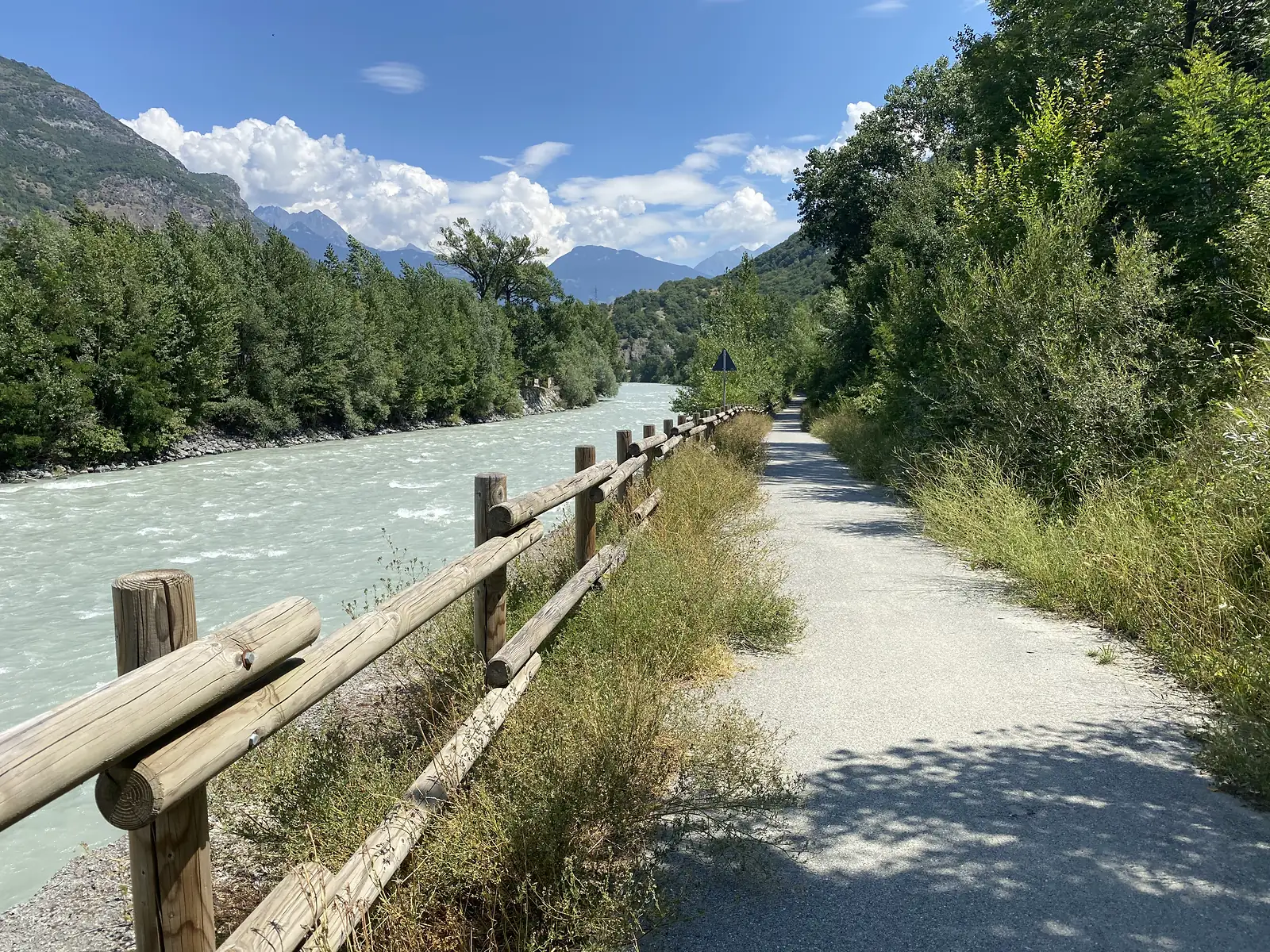 A wide, milky turquoise river flows past a fenced gravel path, wooded hills and distant peaks framing the valley ahead.
