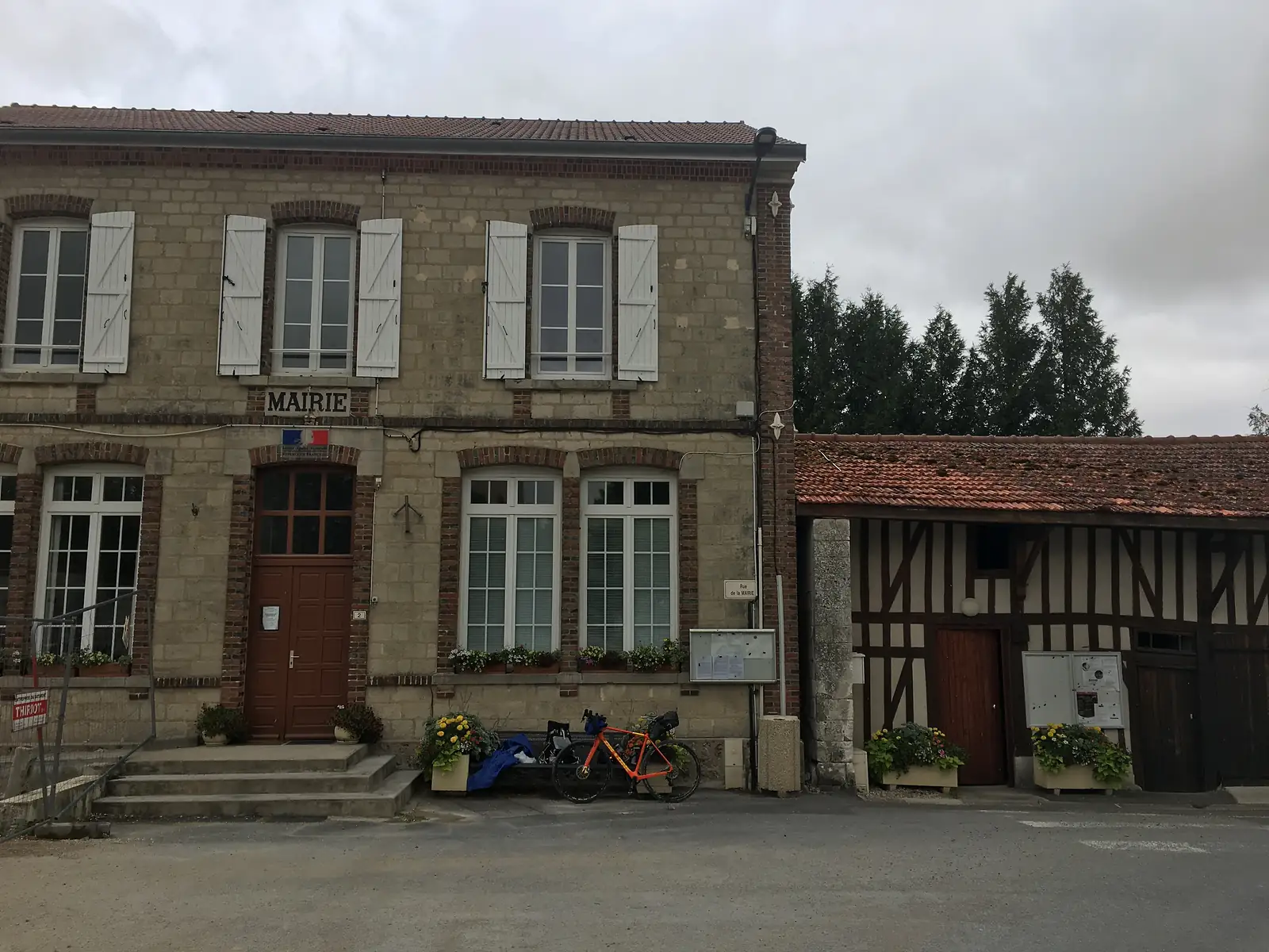 An orange loaded gravel bike leans against a bench outside a village mairie with white-shuttered windows, next to an adjoining half-timbered building.