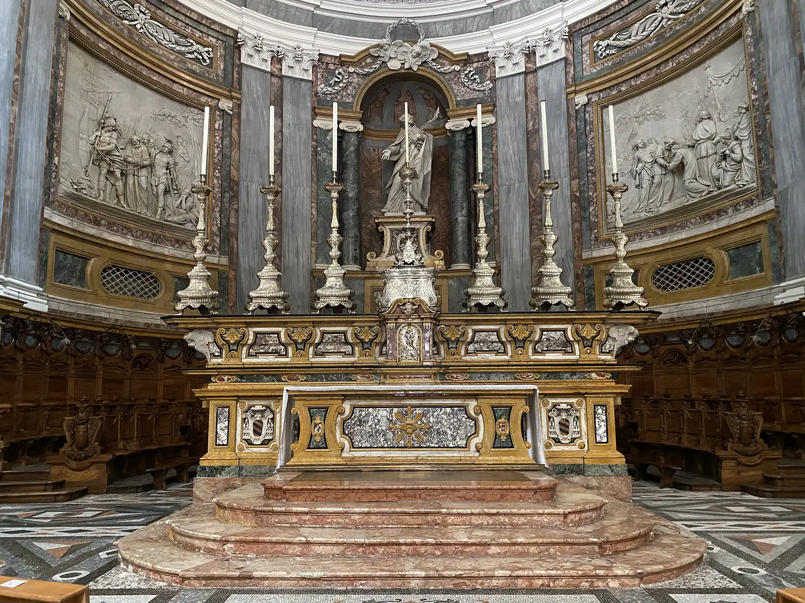 The high altar of Vercelli Cathedral, its marble steps flanked by tall silver candlesticks beneath a niche statue of a bishop saint, with carved relief panels on either side.