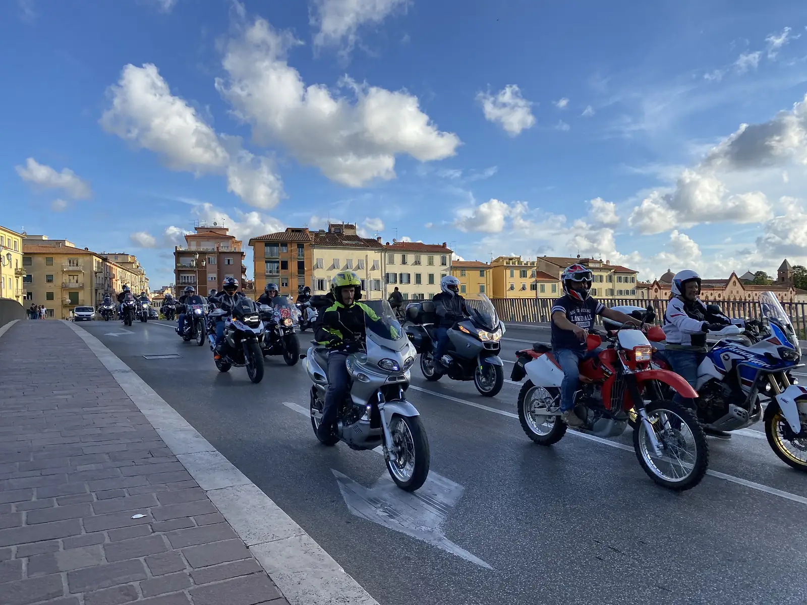 A line of motorcyclists in helmets rides across a stone bridge lined with pastel townhouses, part of a large group ride through the town.