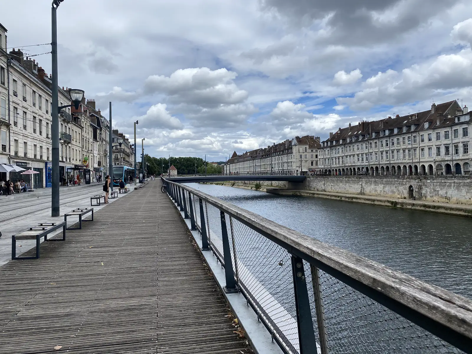 A wooden riverside boardwalk runs alongside the Doubs in Besançon, a tram crossing a stone bridge and terraced townhouses lining the far bank.