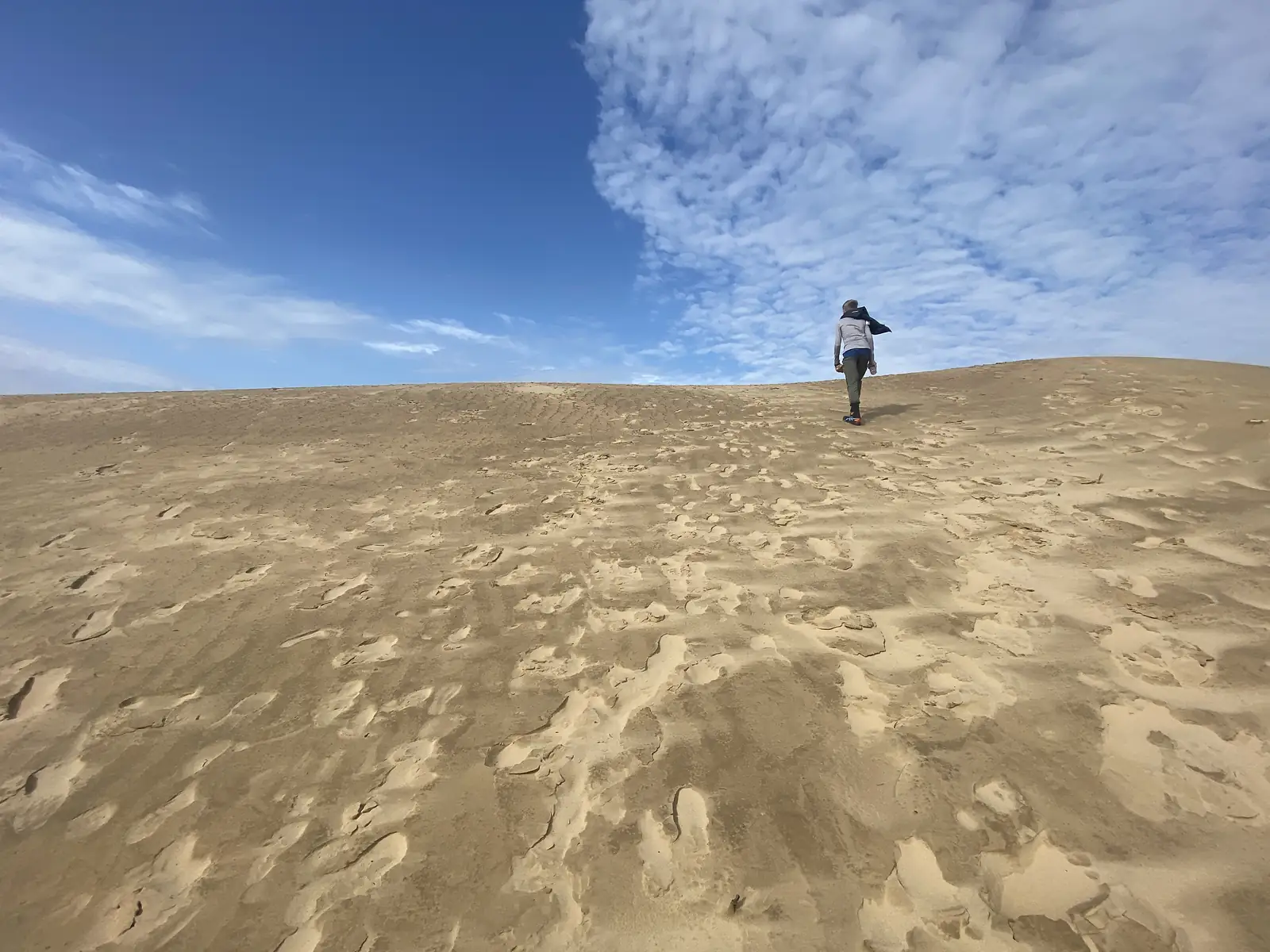 A lone figure walking up a vast bare sand dune under a big blue sky streaked with cloud.