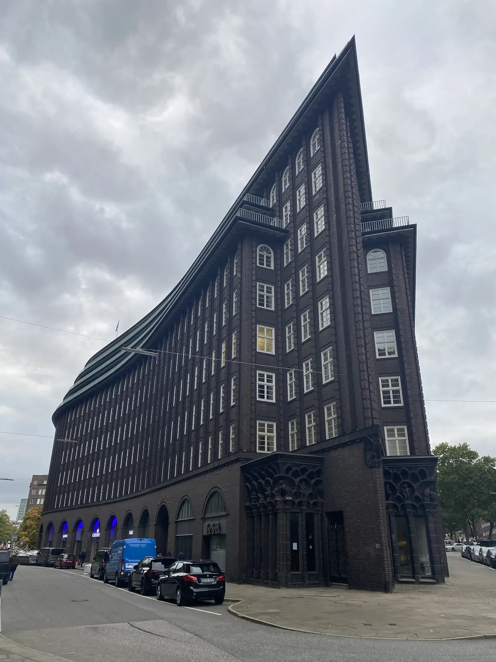 A tall brick office building with a sharply pointed, ship-like prow rises above a street lined with parked cars.