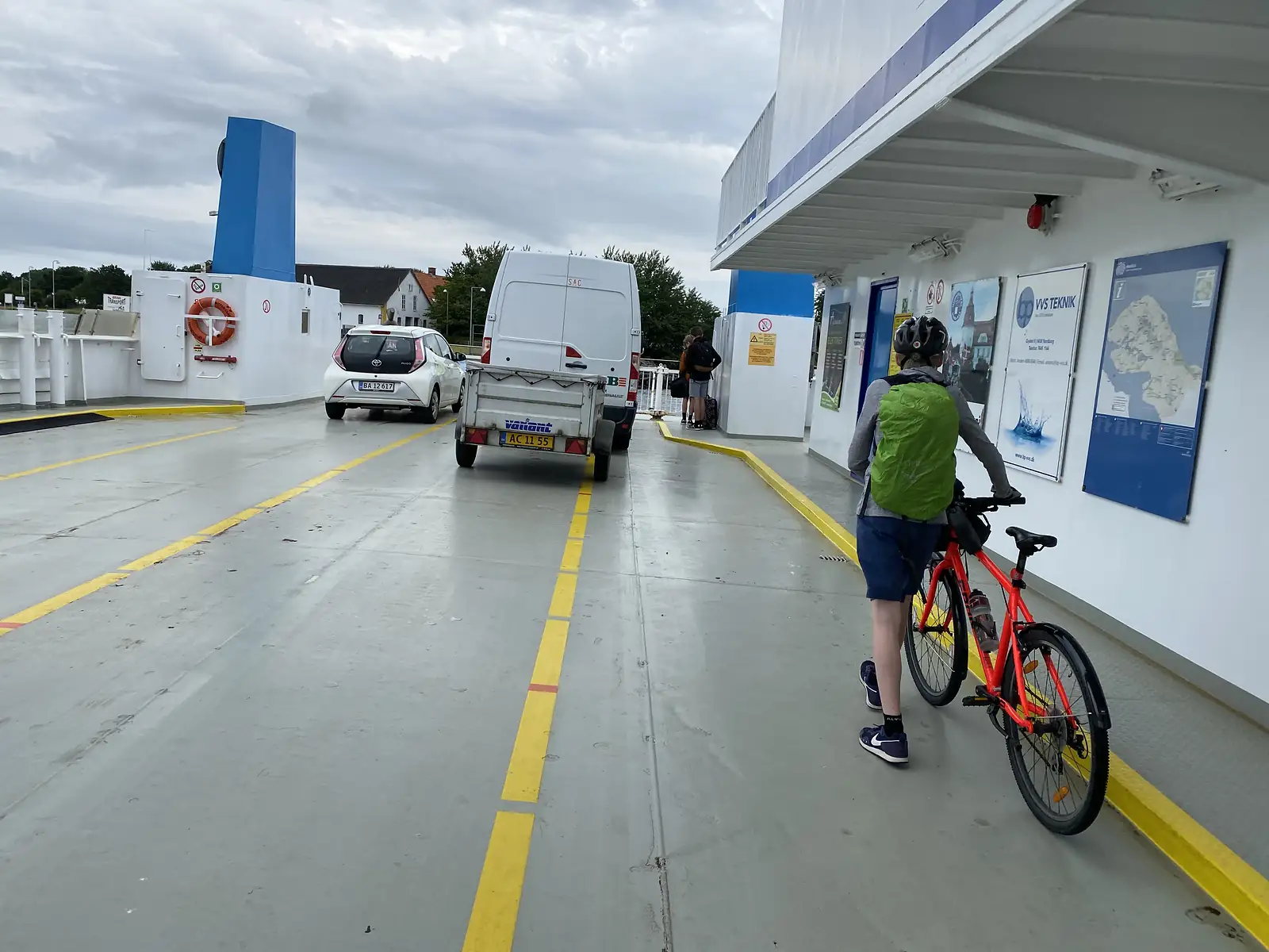 A young cyclist with a green rucksack wheeling a red bike onto a ferry's vehicle deck past a van and trailer.
