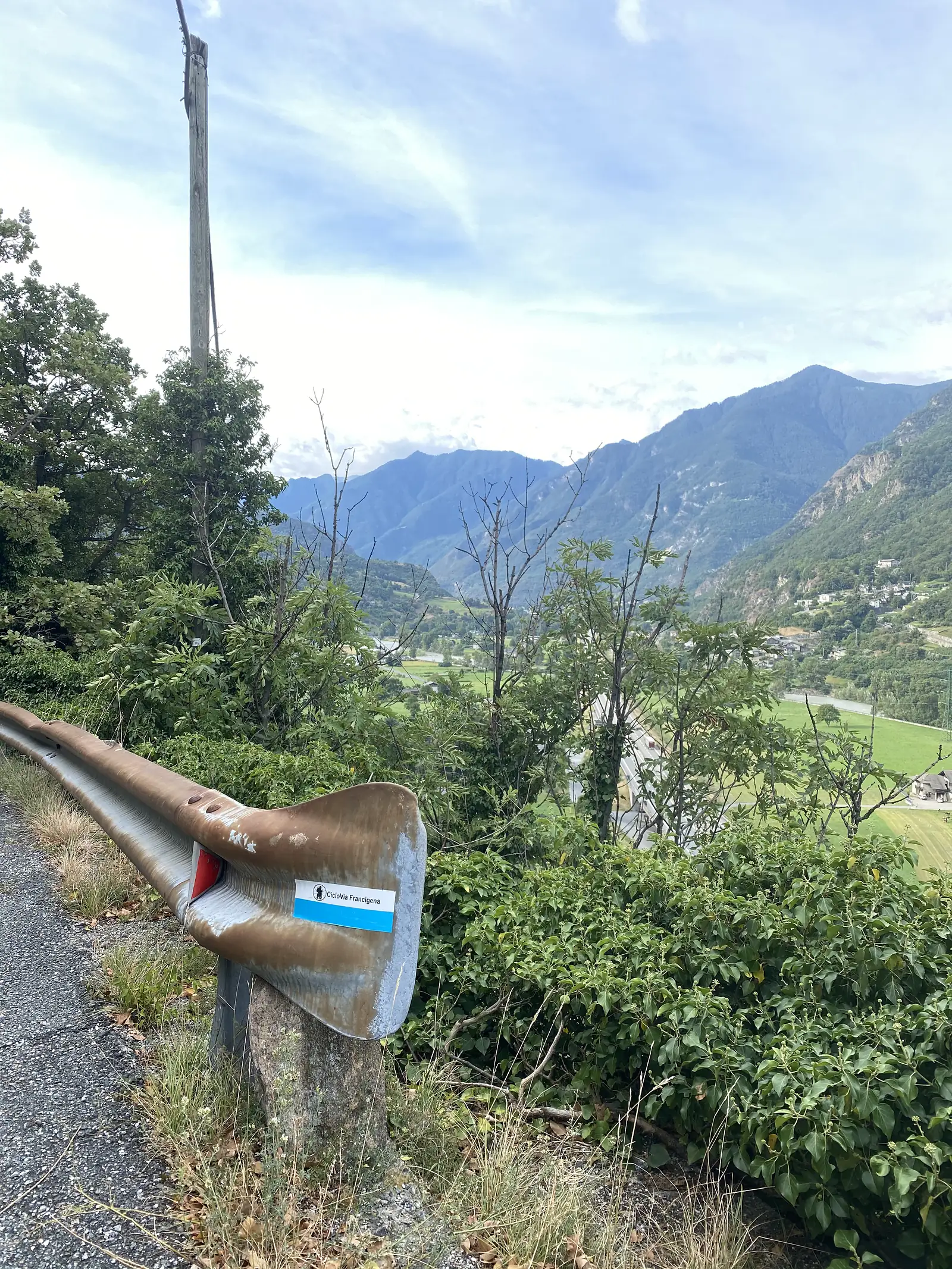 A crash barrier post carrying a blue and white "CicloVia Francigena" route marker sticker, with a wooded valley, river plain and distant mountains behind.
