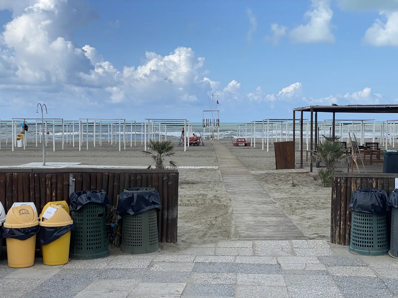 A boardwalk running between recycling bins and rows of shuttered beach umbrellas down to a lifeguard tower and the sea.
