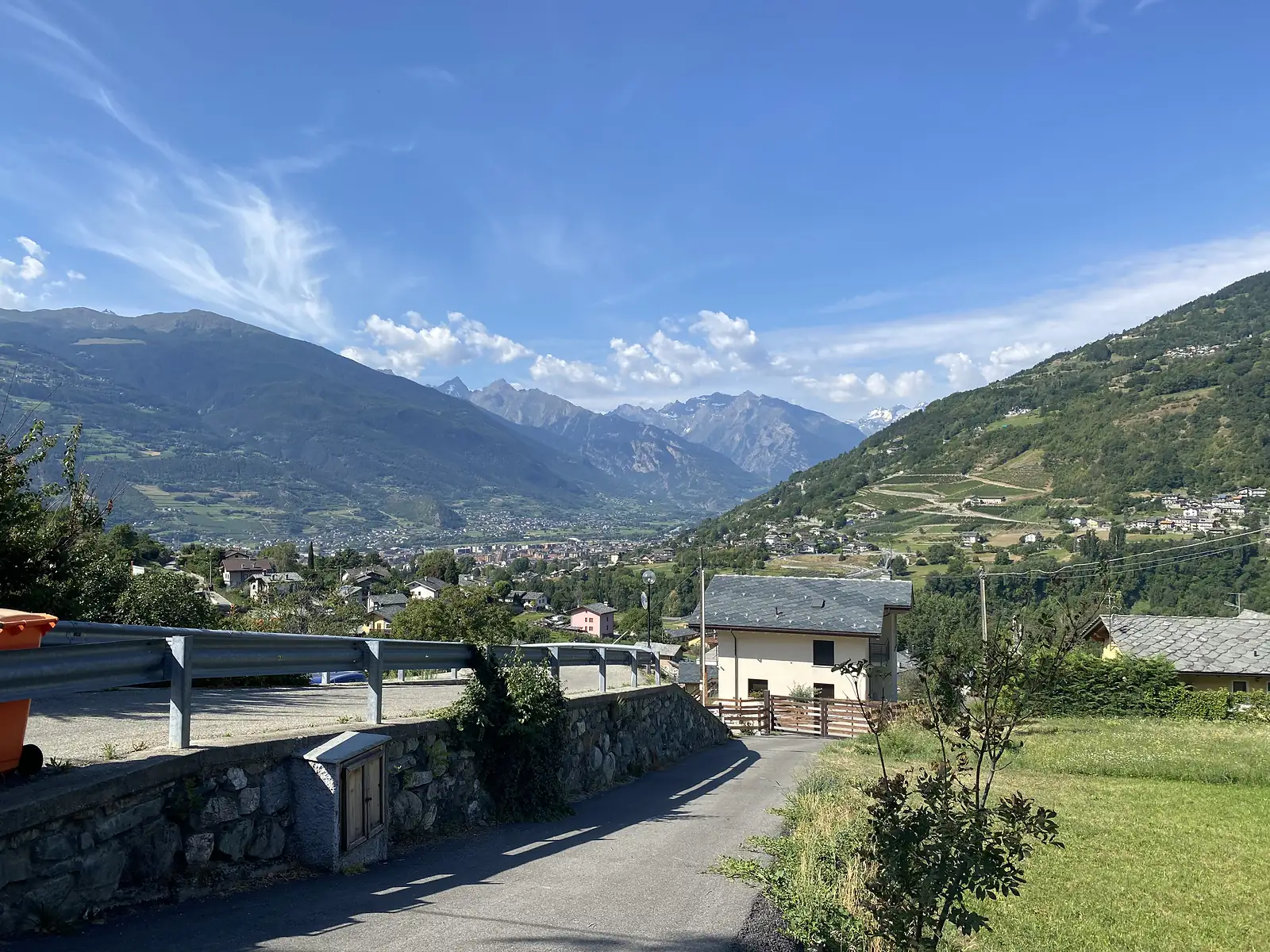 A stone farmhouse and guardrail overlook a wide mountain valley, a town of grey and orange rooftops spread across the valley floor below and a terraced hillside opposite.