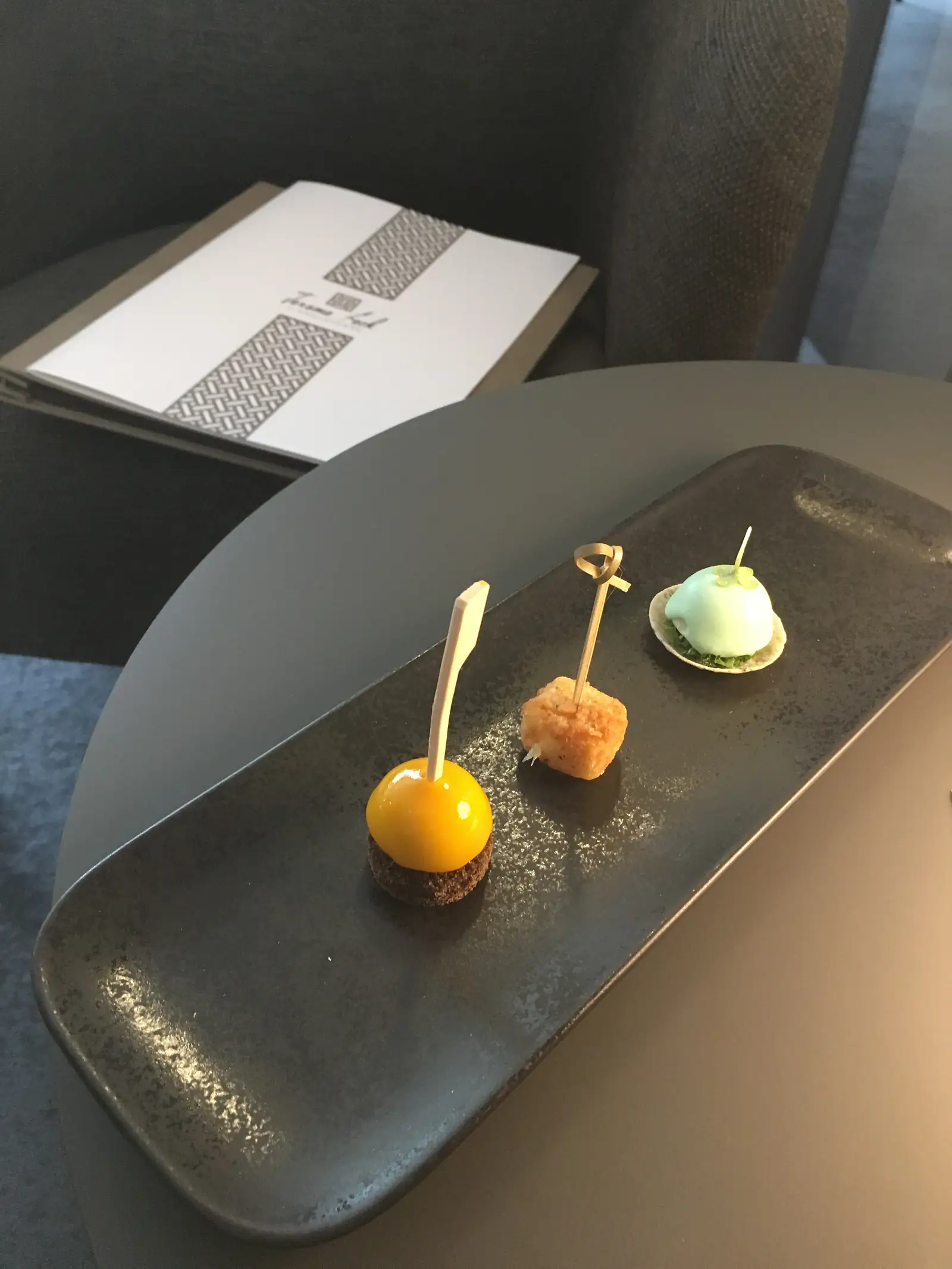 Three delicate amuse-bouches, including a pale green quenelle in a tuile shell and a bright orange sphere on a chocolate-dusted base, arranged along a dark speckled plate.