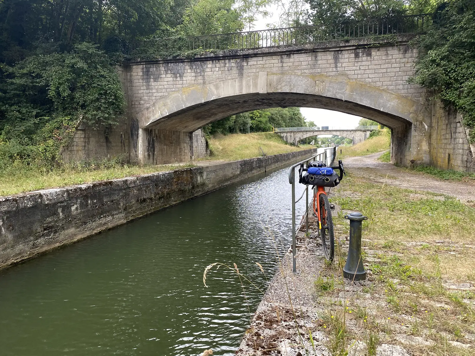 A loaded orange bikepacking bike leans against canal railings beneath a stone road bridge, with a second bridge visible further along the waterway.