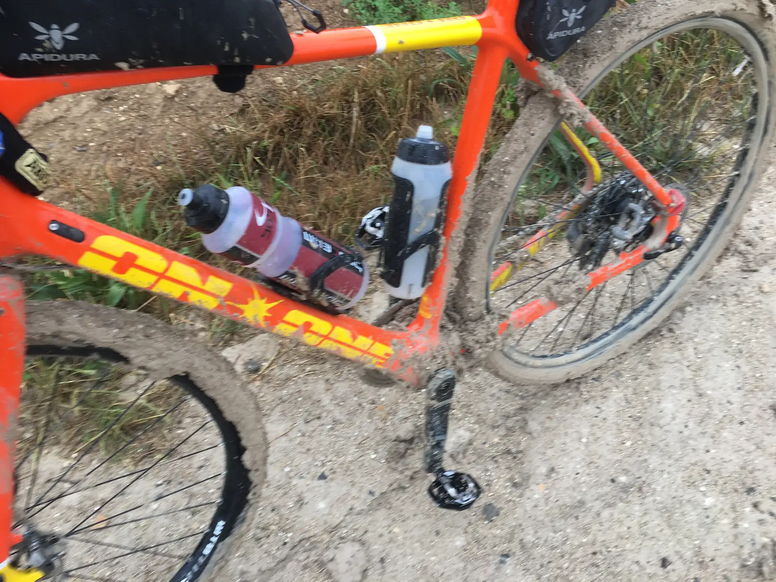 Close-up of a thickly mud-caked bicycle frame and rear wheel, with two water bottles wedged between the tubes and dry grass verge behind.