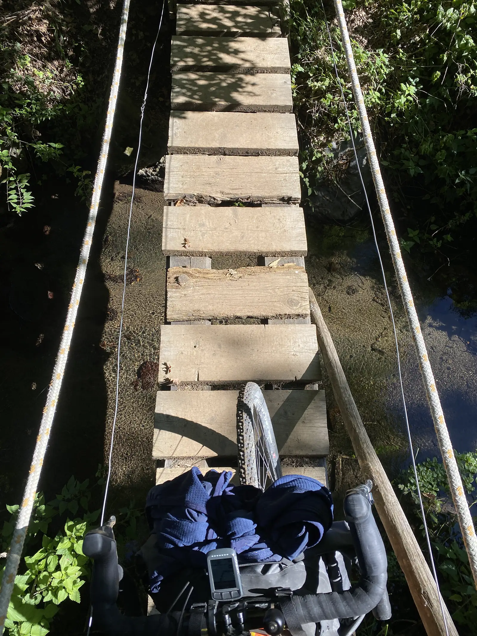 A loaded bike is wheeled across a narrow wooden-plank footbridge with rope handrails over a clear stream.