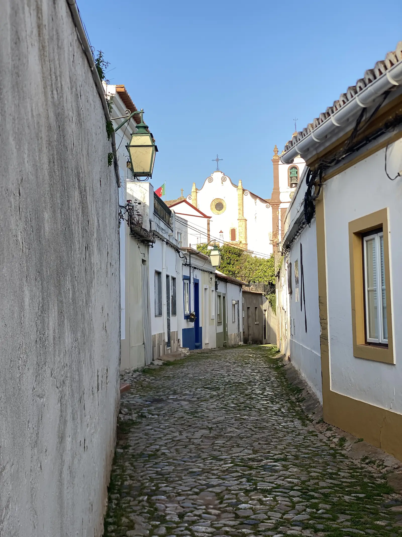 Narrow cobbled lane running between low whitewashed houses towards a white church with a round rose window, scrolled gable and bell tower, a wrought-iron lantern projecting from the wall in the foreground.