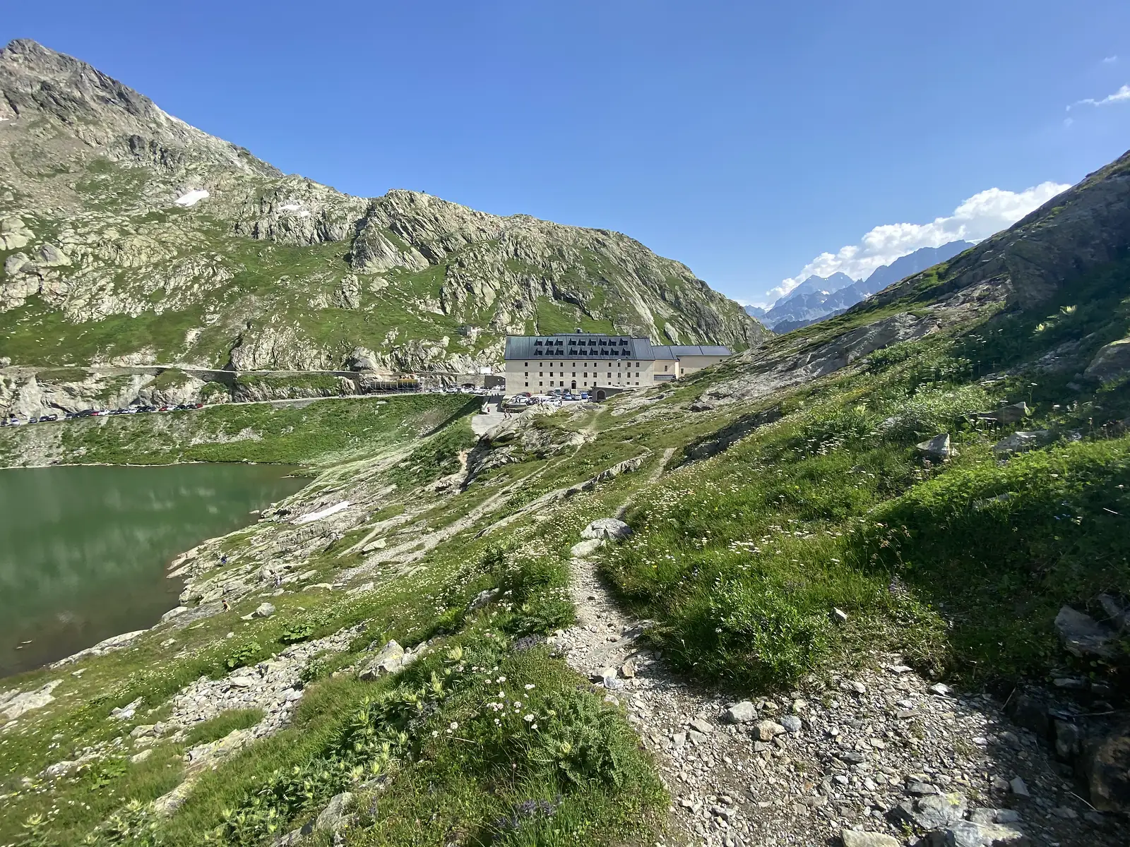 The hospice building above a green mountain lake, with a rocky path leading down towards the water and snow-streaked peaks in the distance.