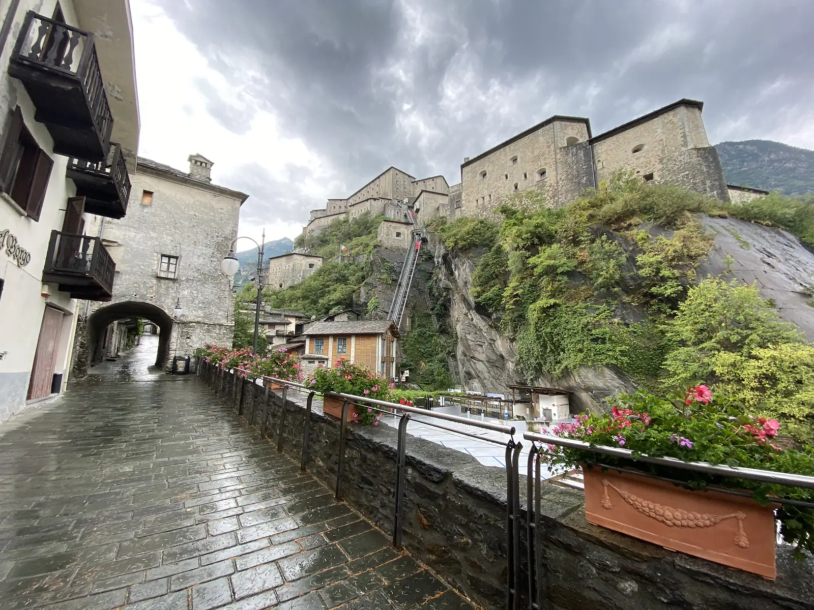 A cobbled lane through the village of Bard leading past flower-lined railings towards a funicular track climbing the cliff face to the fort above.