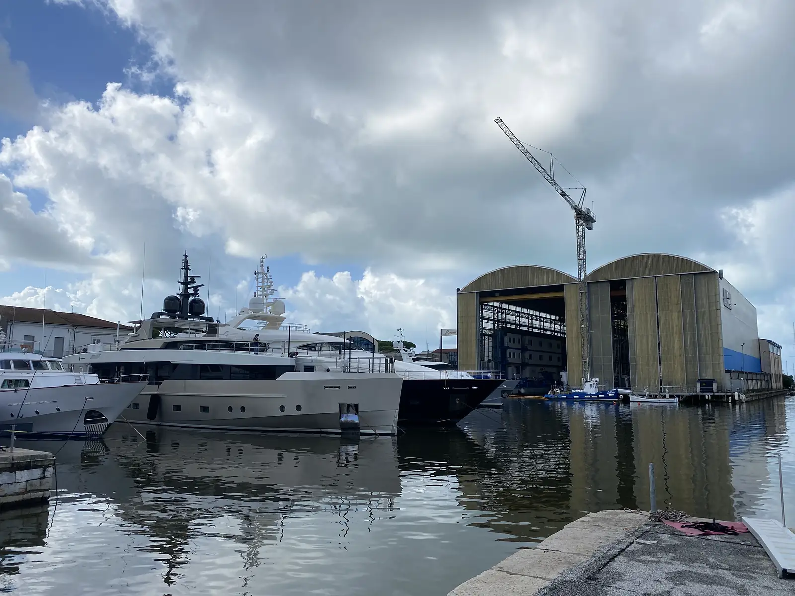 Superyachts moored in a marina beside a shipyard with a tall crane and arched steel sheds under a cloudy sky.