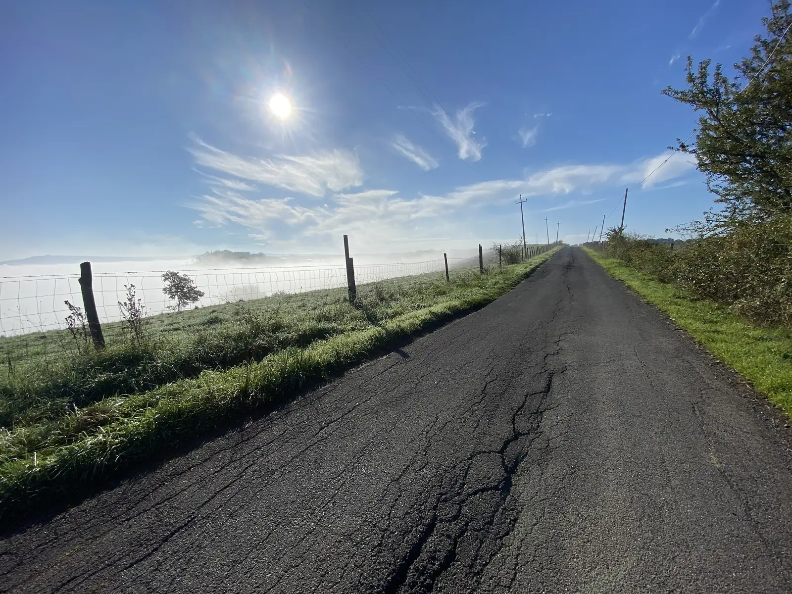 A cracked tarmac lane follows a ridge above a valley of low cloud, with a wire fence and power poles running alongside.