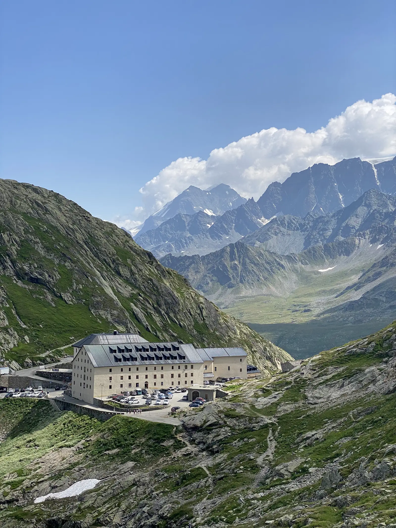 The hospice's shuttered stone facade and dormer roof set against a backdrop of sharp, snow-streaked summits and a broad glacial valley.