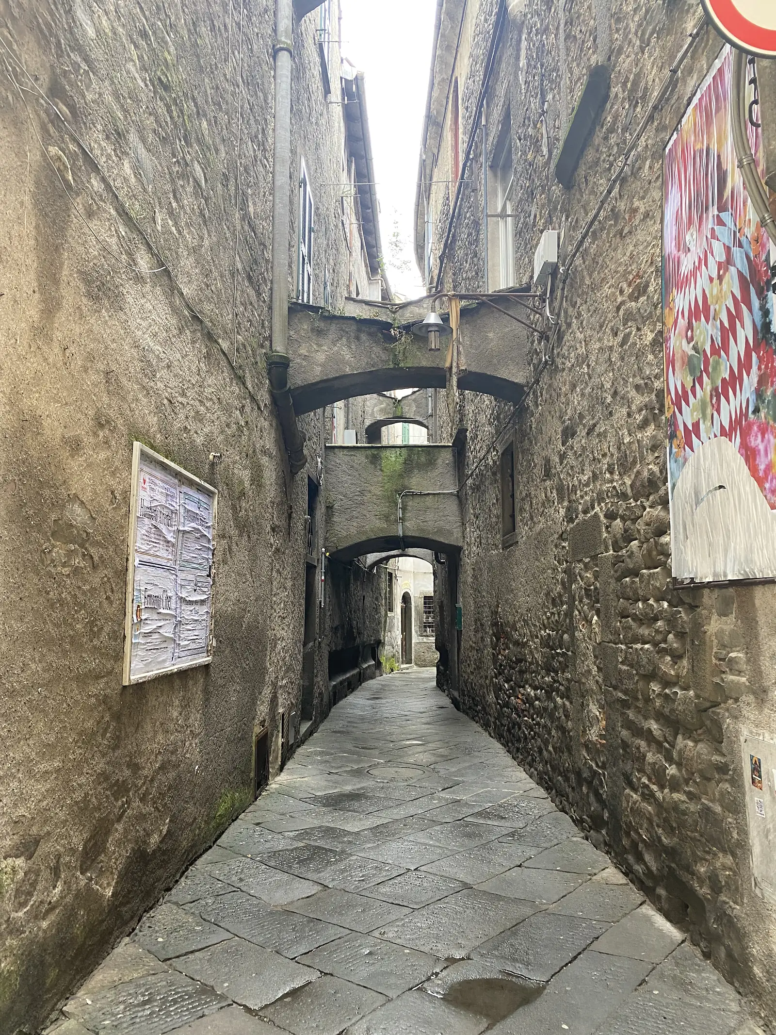 A vaulted stone passageway runs beneath a village building, its walls hung with faded notices and a map board propped on the ground.