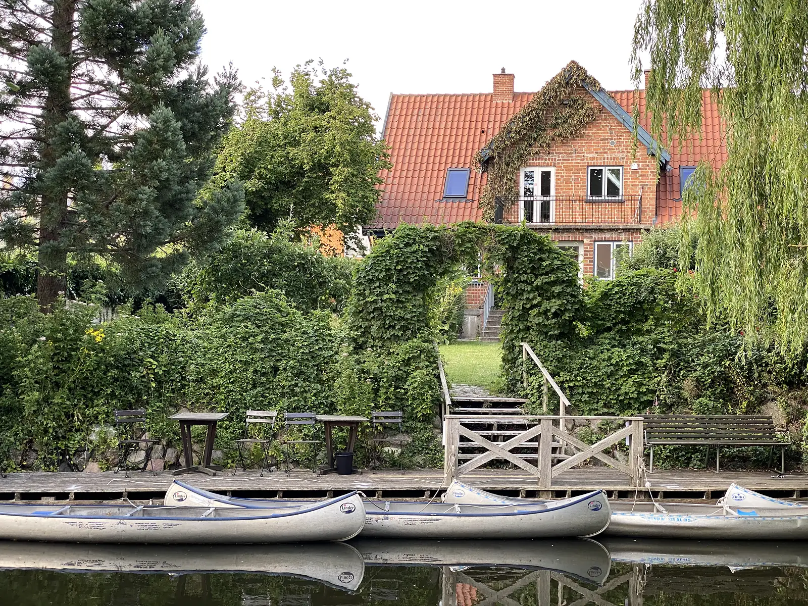 Aluminium canoes moored along a wooden staging below a garden of clipped hedges and an ivy arch, with a red-brick house behind.