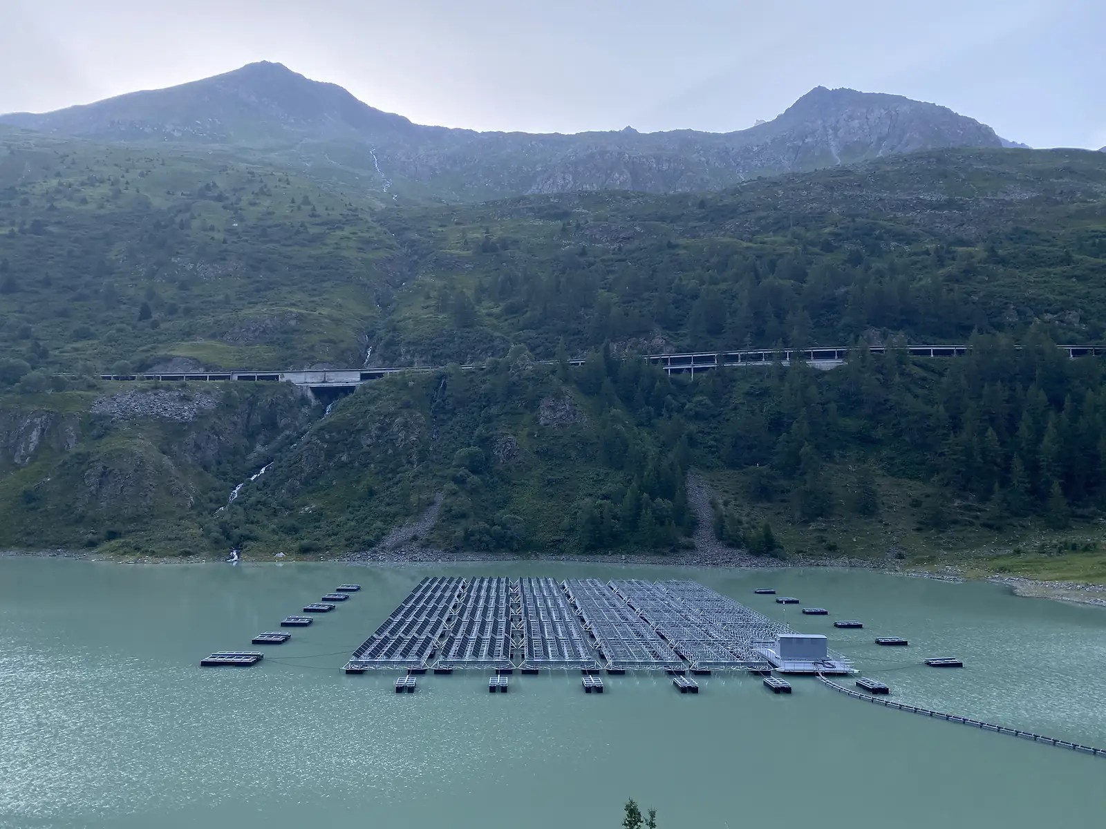 A large array of floating solar panels covers part of a turquoise alpine reservoir, with a road bridge and forested slopes behind.