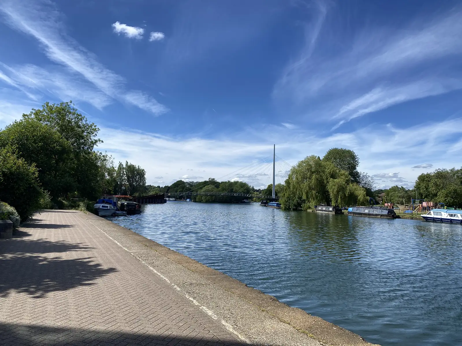 A wide river with moored narrowboats and cruisers along the far bank and a white cable-stayed footbridge mast in the distance.