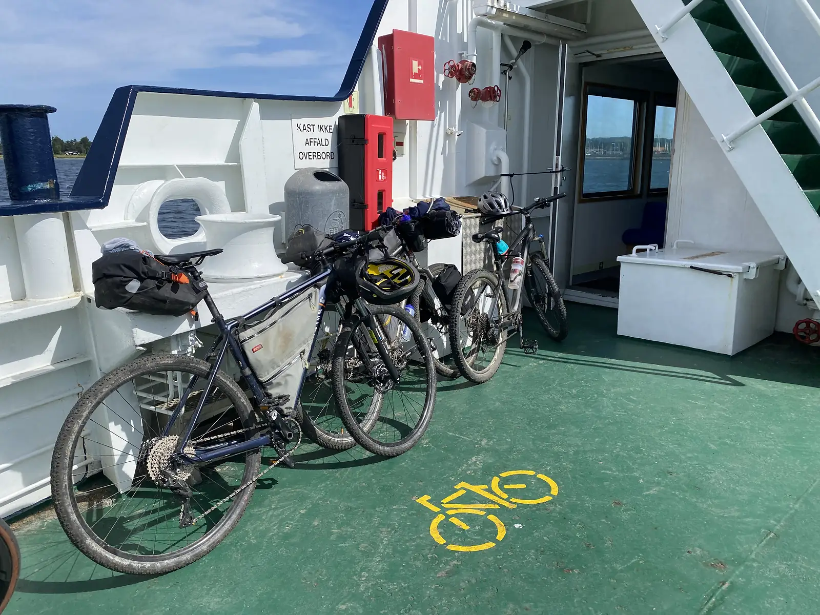 Three loaded bikepacking bikes against the rail of a ferry's green car deck, beside a painted yellow bicycle symbol.