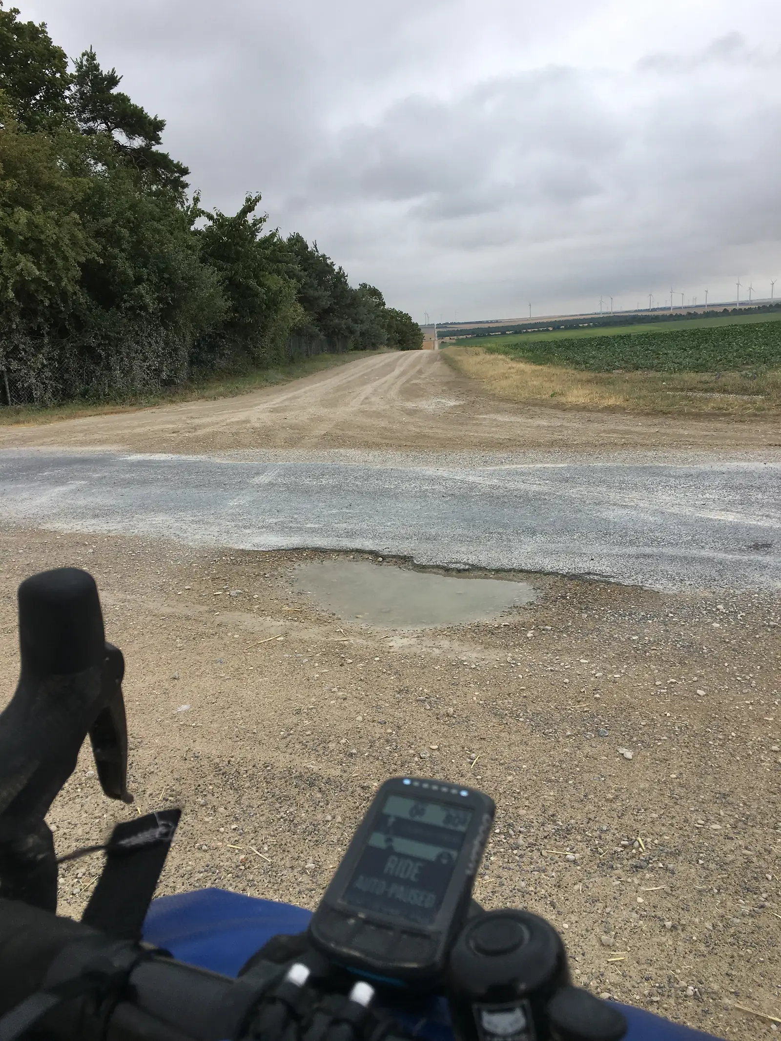 A pale gravel track cuts through open farmland towards distant wind turbines under a heavy grey sky, seen from behind a bicycle's handlebars.
