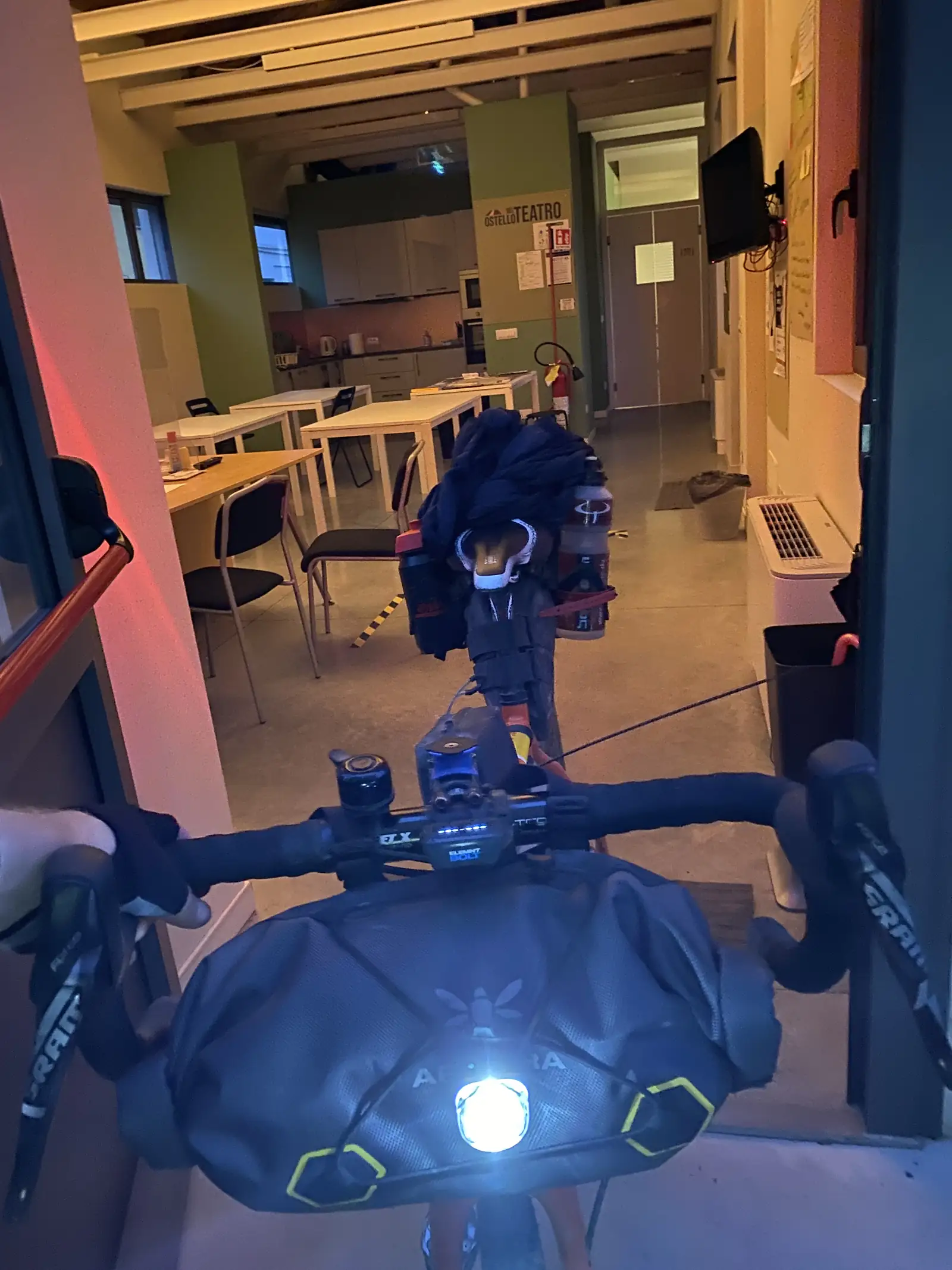 A loaded touring bicycle with a lit front light standing in the dim lobby of the Ostello Teatro hostel, chairs and pigeonholes stacked behind it.