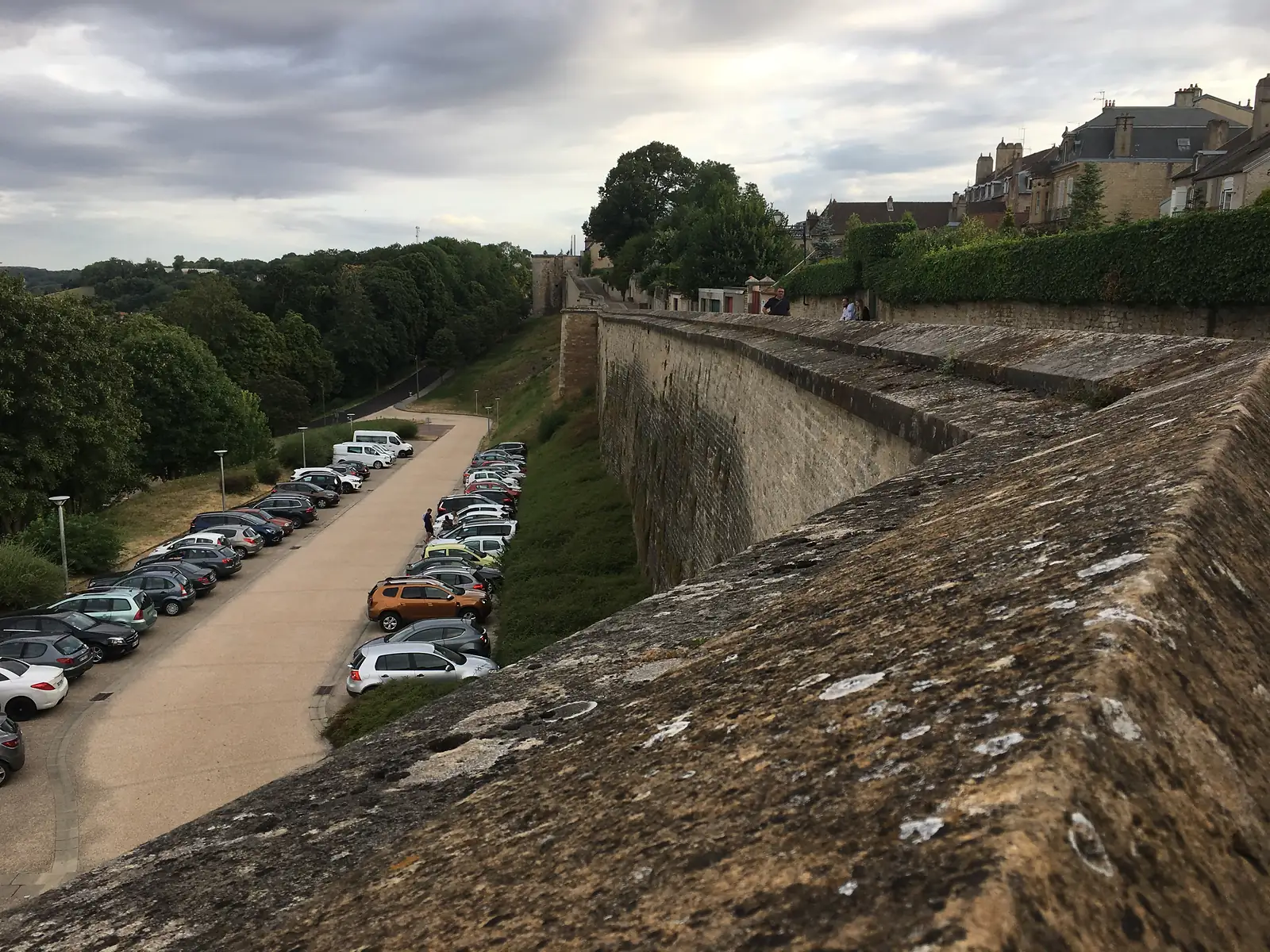 The Langres ramparts walk along a high stone wall above a car park and wooded slope, with the old town's rooftops visible ahead under a dramatic evening sky.