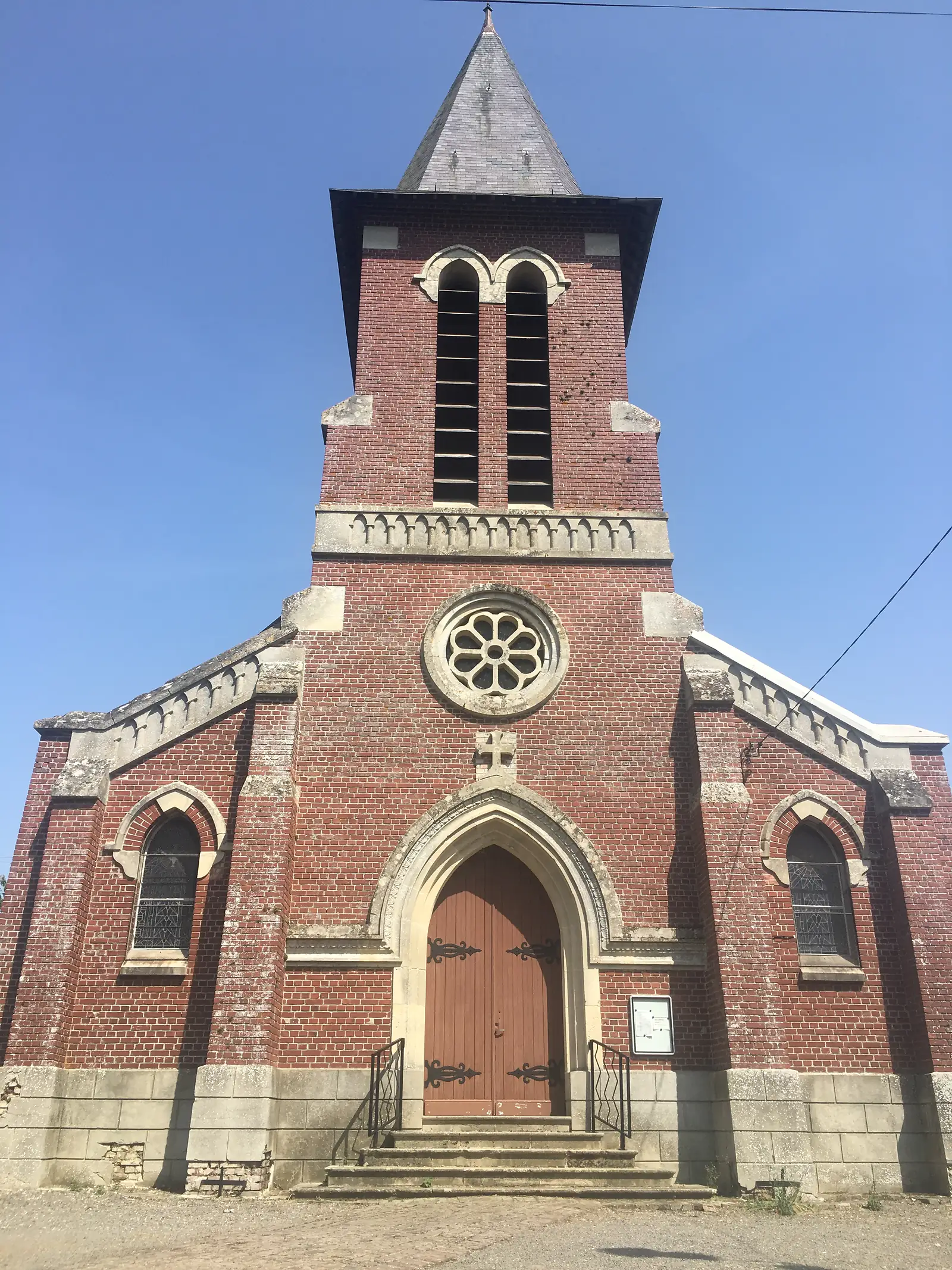 A red-brick village church with a stone rose window and a tall slated spire, set against a clear blue sky.