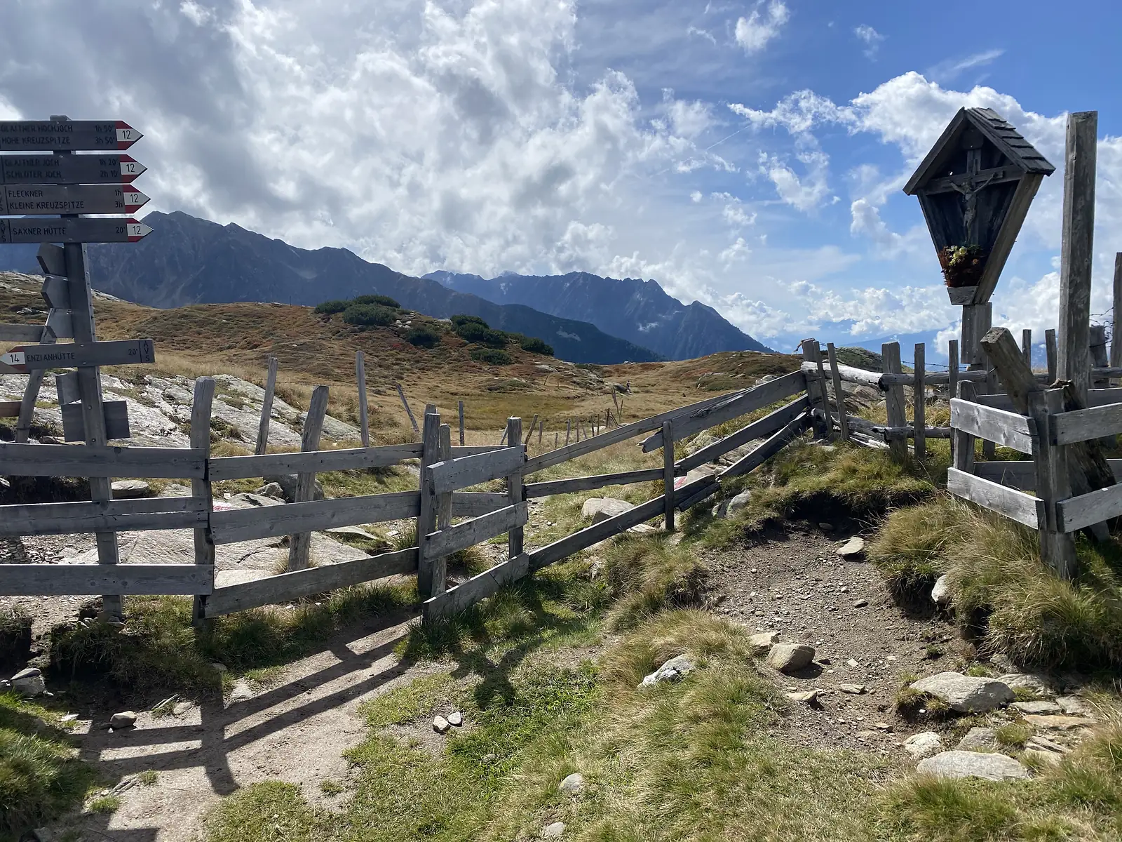 A hikers' fingerpost pointing to Glaitner Hochjoch, Hohe Kreuzspitze, Fleckner and the Saxner Hütte stands beside a timber wayside shrine where a path crosses a split-rail fence on an open mountain pass.