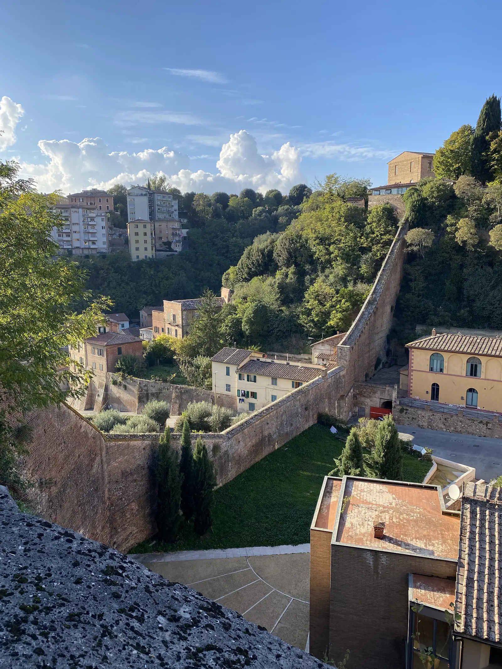 A stone-walled path running along a hillside through a Tuscan town, lined with cypress trees and terracotta-roofed houses beneath a partly cloudy sky.